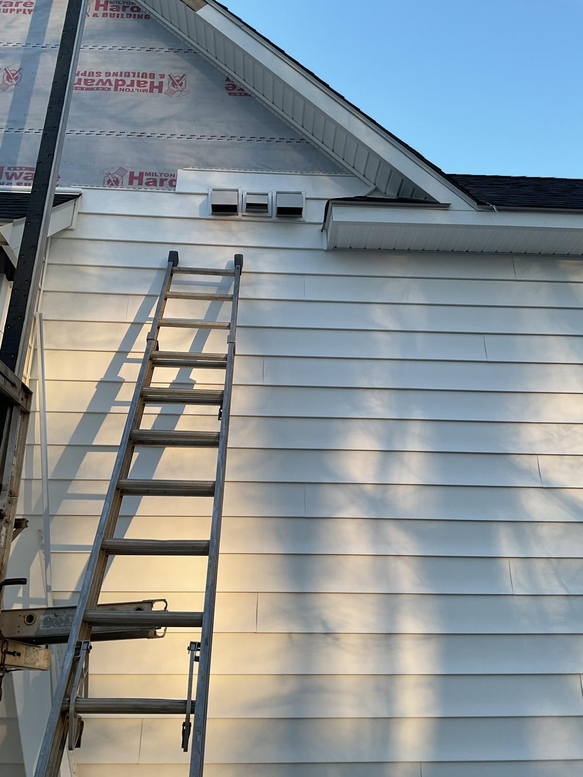 A ladder leans against the side of a house with white horizontal siding and exposed weather-resistant house wrap above.