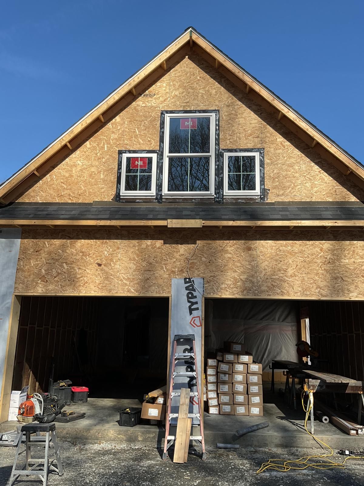 A construction site showing a house frame with three windows installed in the gable wall and a ladder in the foreground.