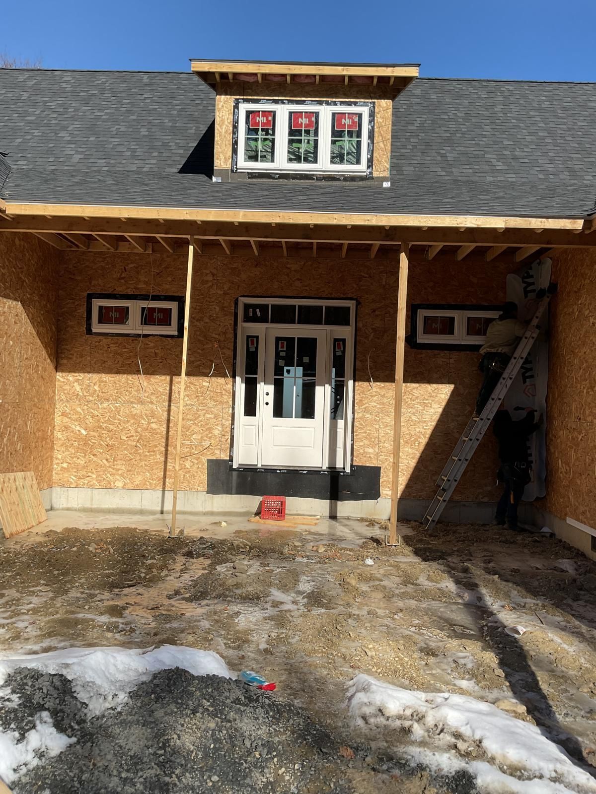 A house under construction with a front door, two small windows, a dormer, and a ladder leaning against the exterior.