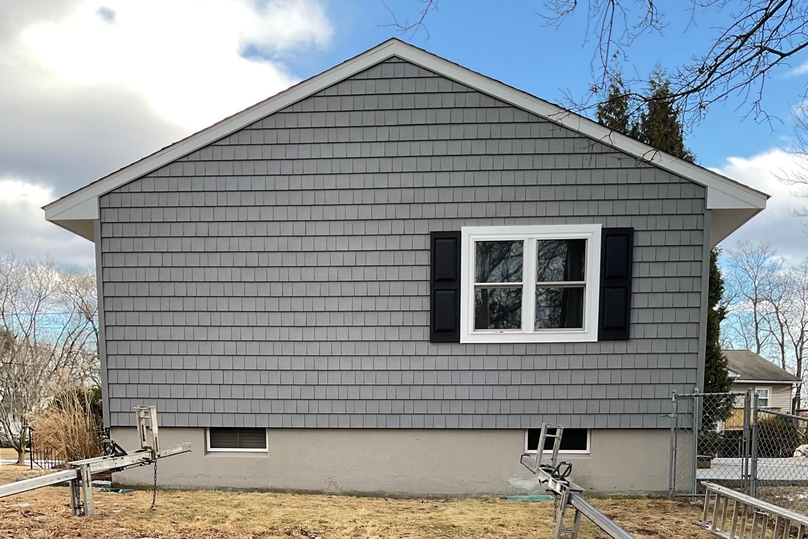 Side view of a house with grey shake siding, a white-framed window with black shutters, and a concrete foundation.