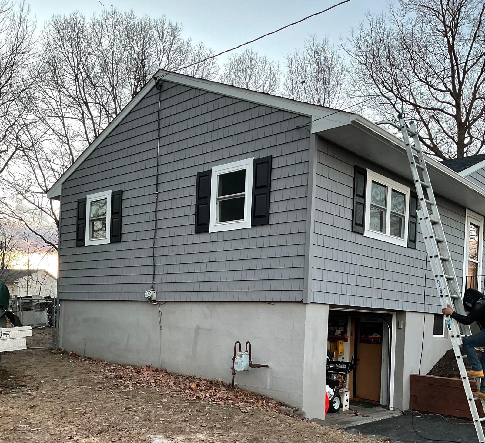 A person climbs an extension ladder leaning against the side of a gray-sided house with a concrete foundation.