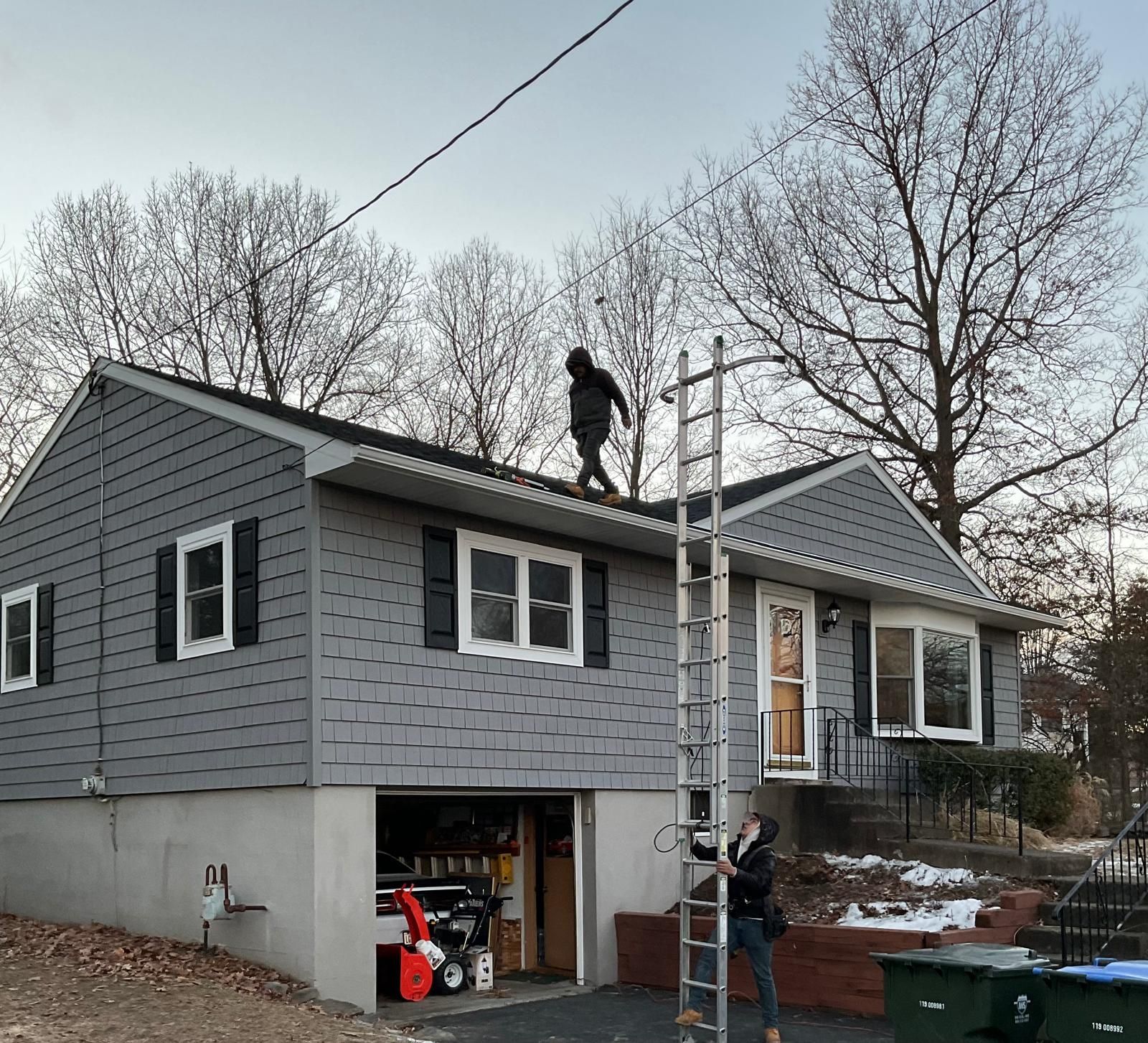 A person climbs a long ladder leaning against a gray-sided house, while another person stands on the roof.