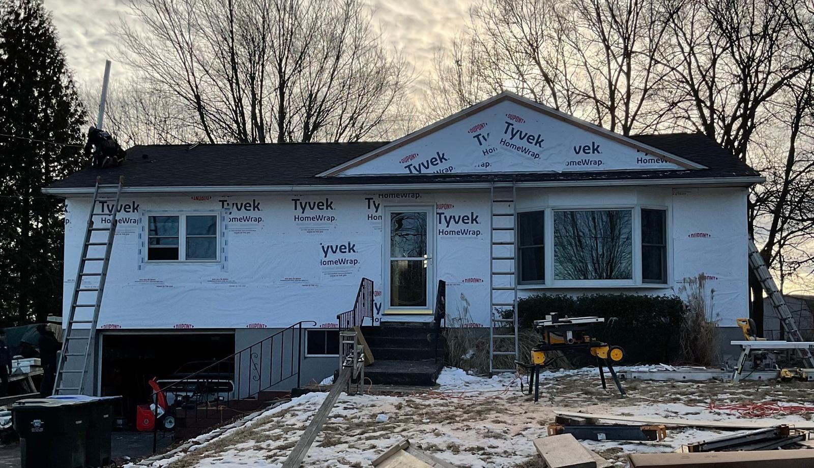 A house undergoing exterior renovations with Tyvek house wrap, ladders, and equipment, set against trees in winter.