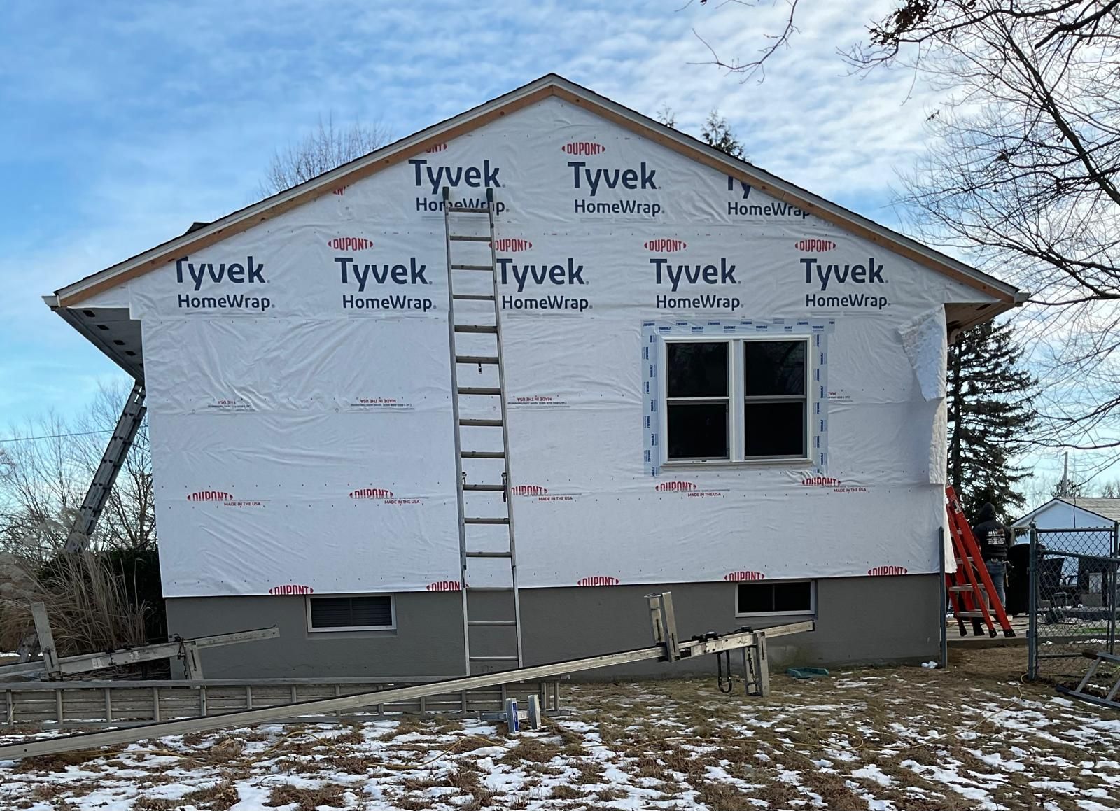 A house exterior covered in white Tyvek wrap, featuring a window, a leaning ladder, and a gray concrete foundation.