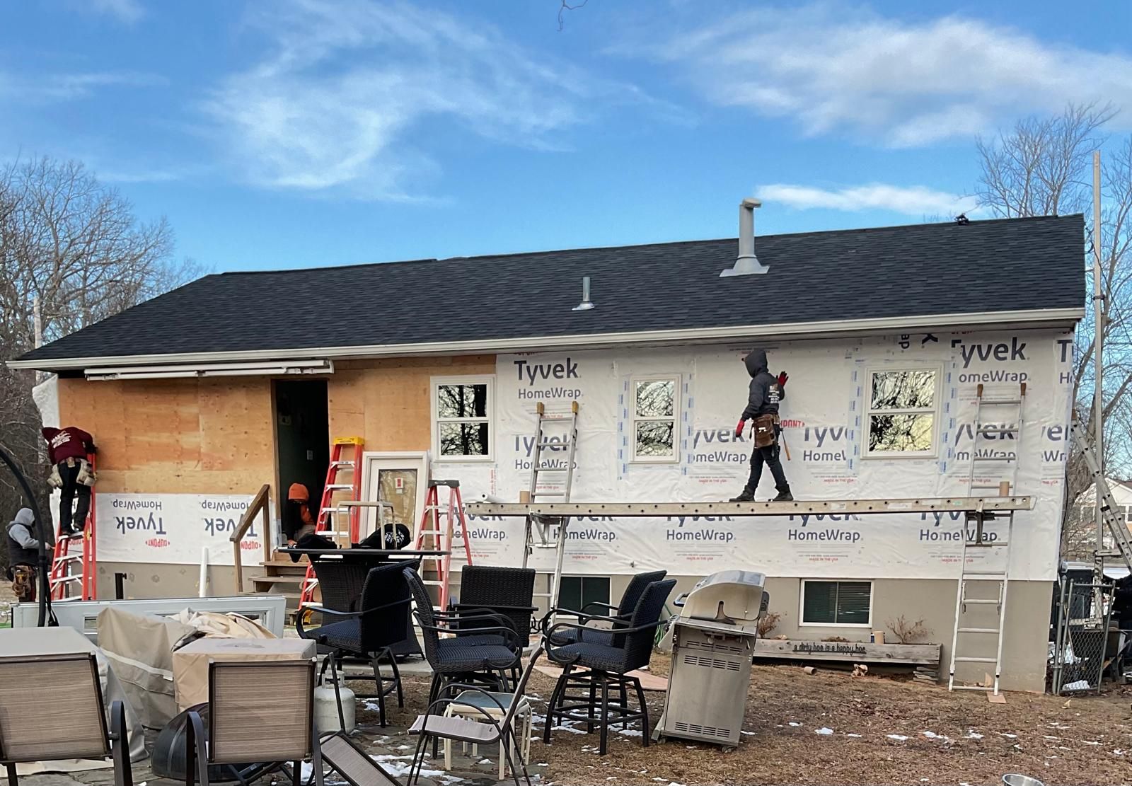 Two construction workers perform exterior renovations on a house, installing white Tyvek siding and new windows.