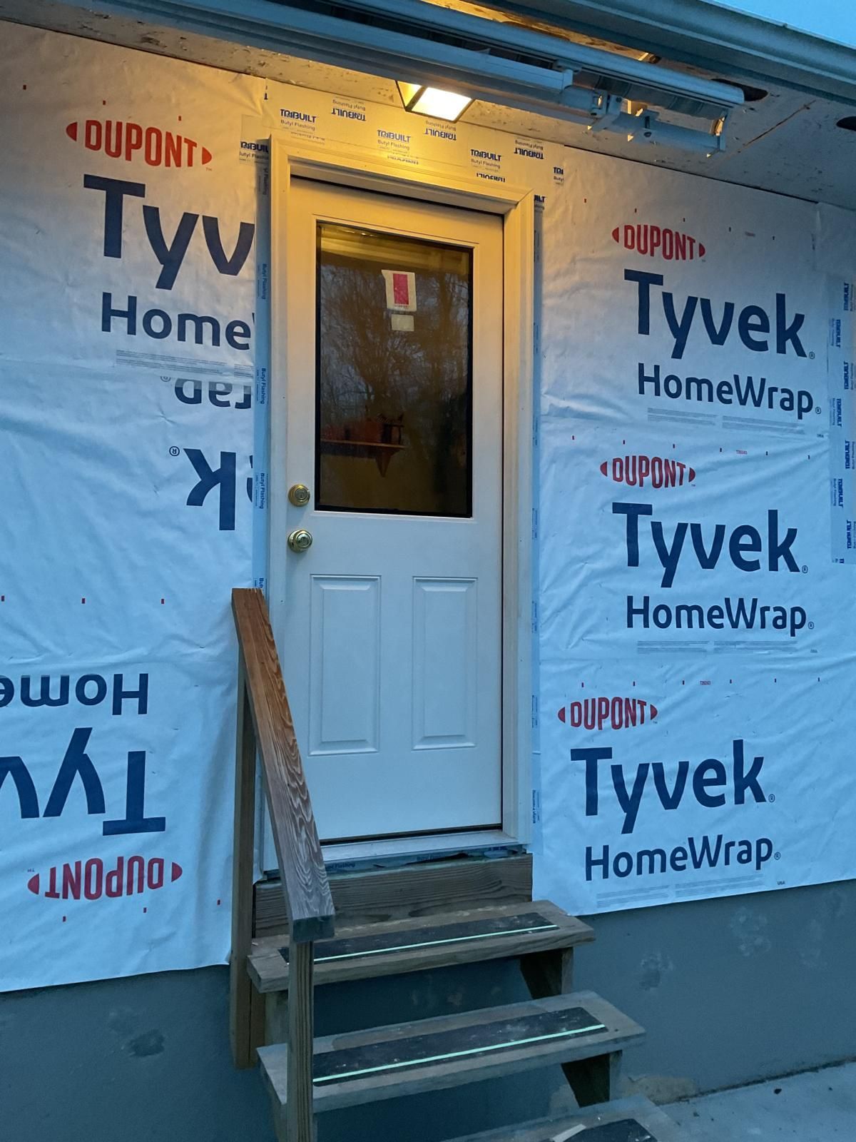 An exterior door with a window and small wooden steps, surrounded by unfinished white Tyvek home wrap siding.