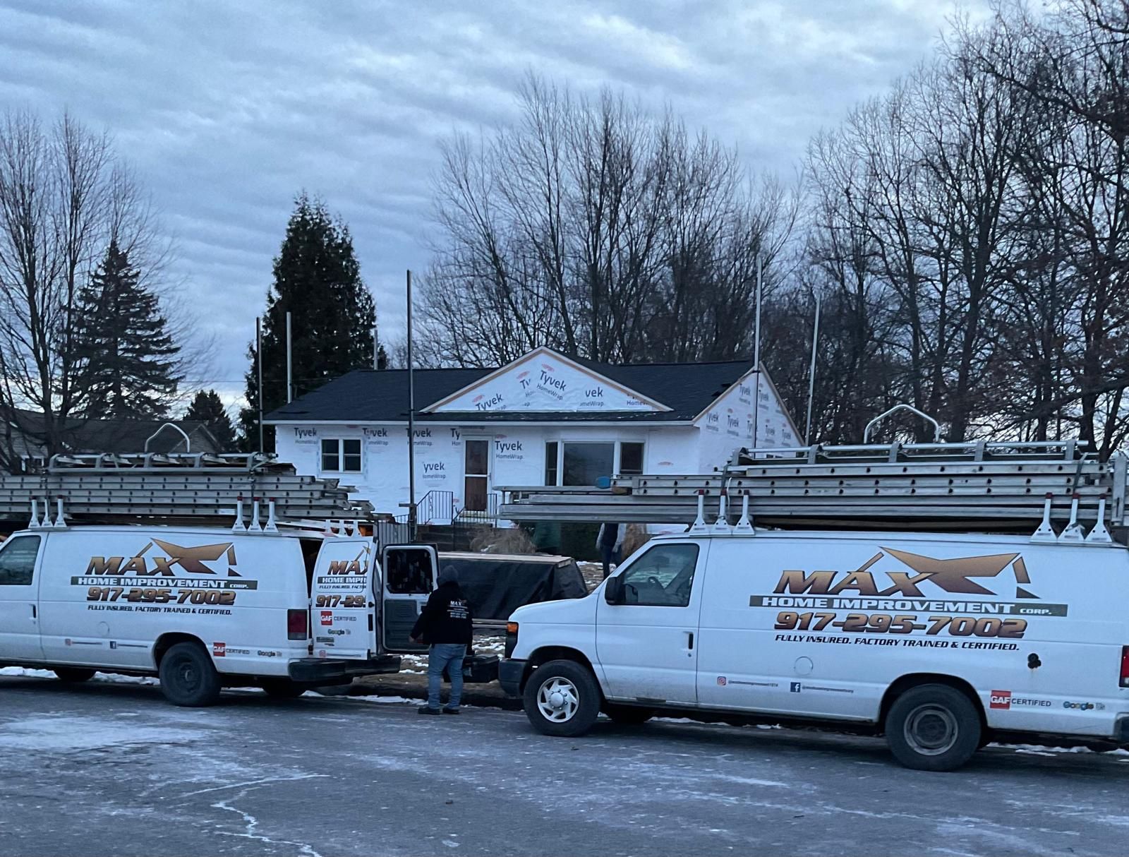Two white Max Roofing vans parked in front of a house under construction during a snowy winter day.