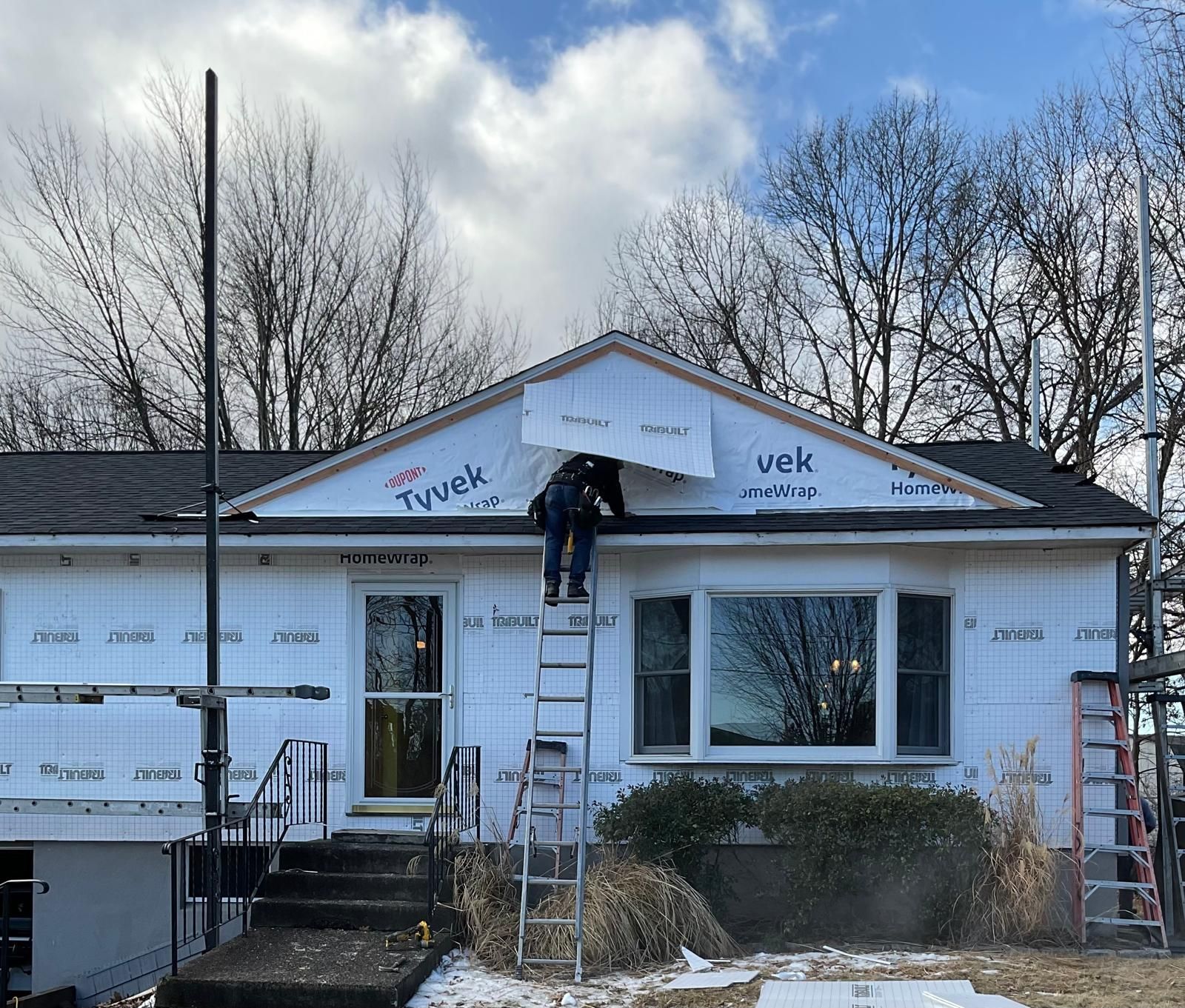 A person on a ladder repairs the gable end of a house covered in white Tyvek weather-resistant building wrap.
