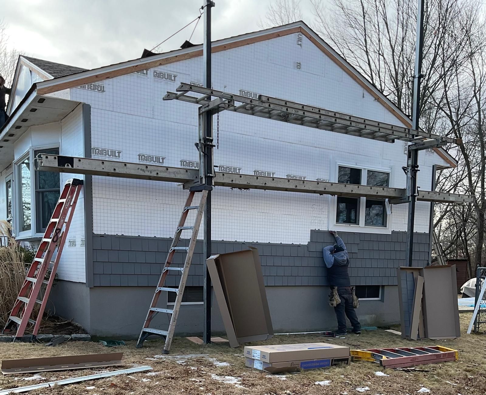 A worker installs grey siding on the exterior of a house under renovation, supported by scaffolding and ladders.