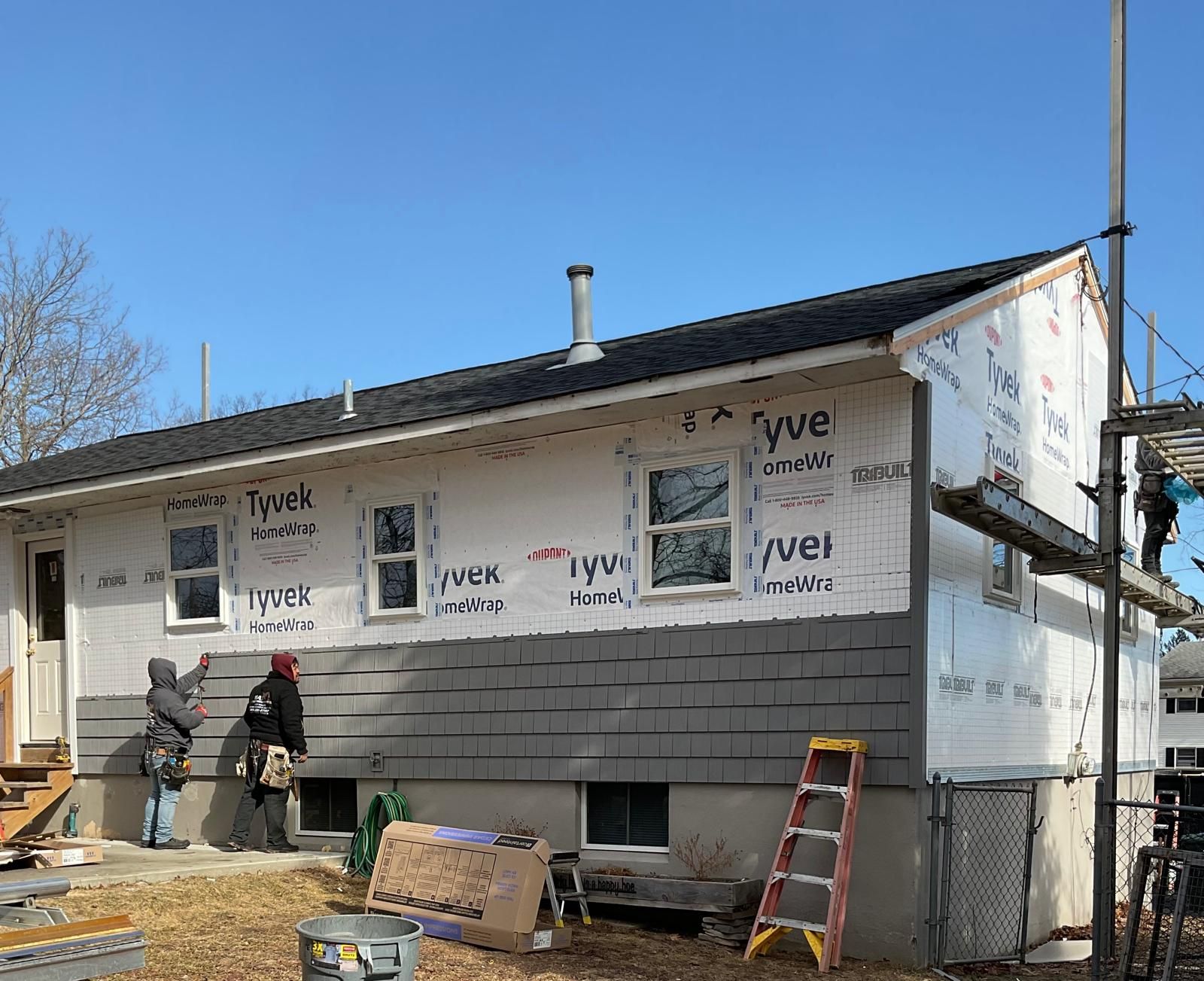 Two workers install gray exterior siding on the side of a white house wrapped in Tyvek moisture barrier.