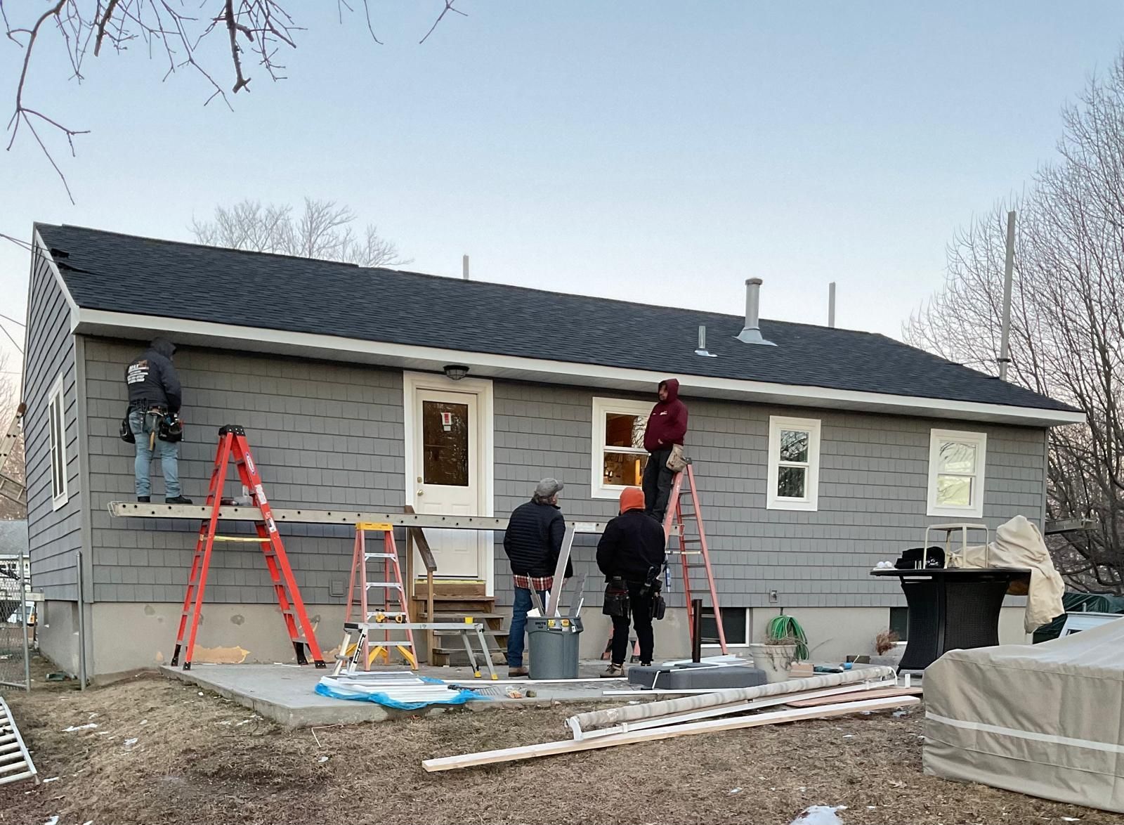 Four workers in casual clothing install grey siding on the side of a house using ladders and scaffolding.