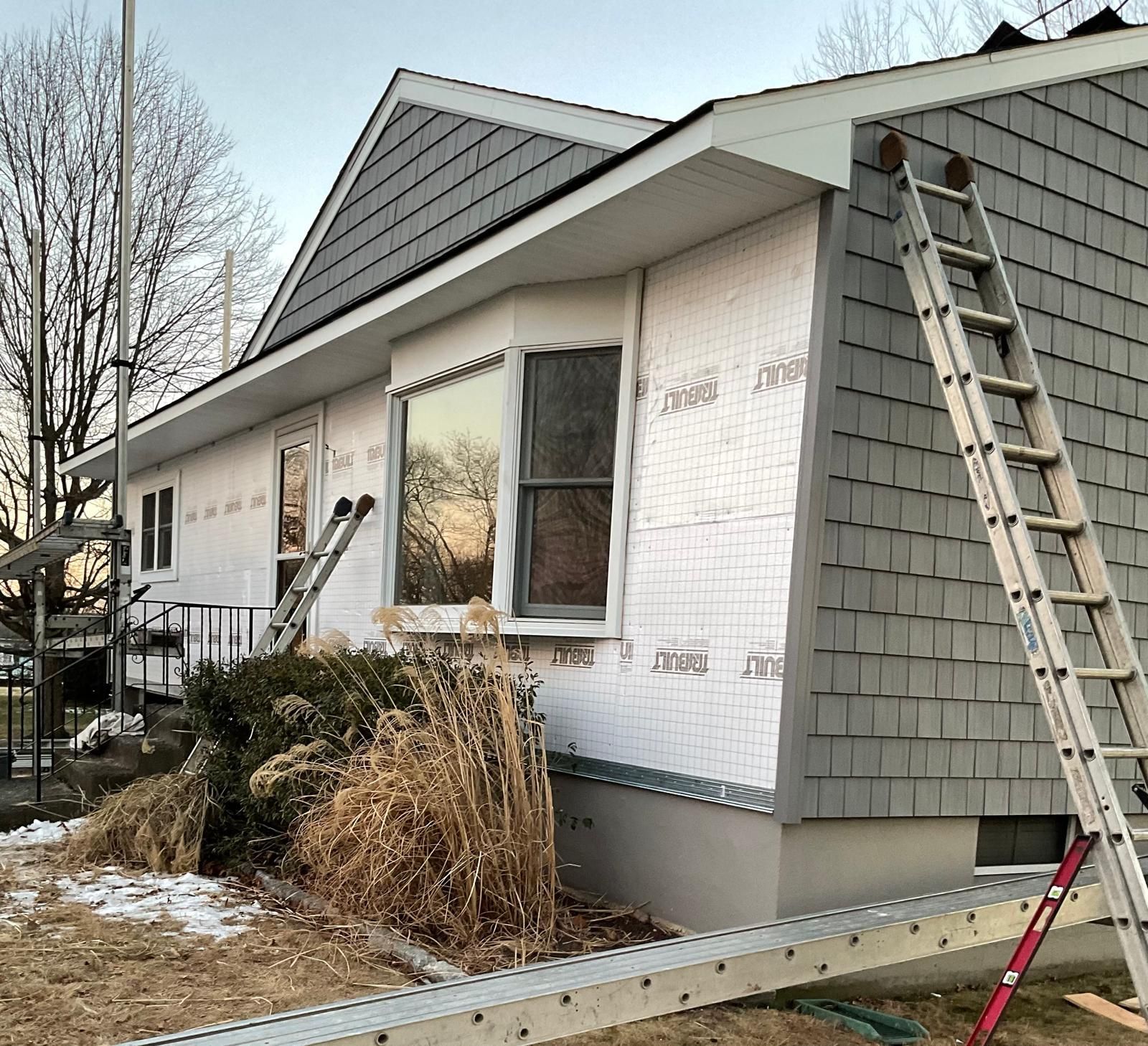 A house under renovation with partially installed gray shingles, white protective house wrap, and two extension ladders.