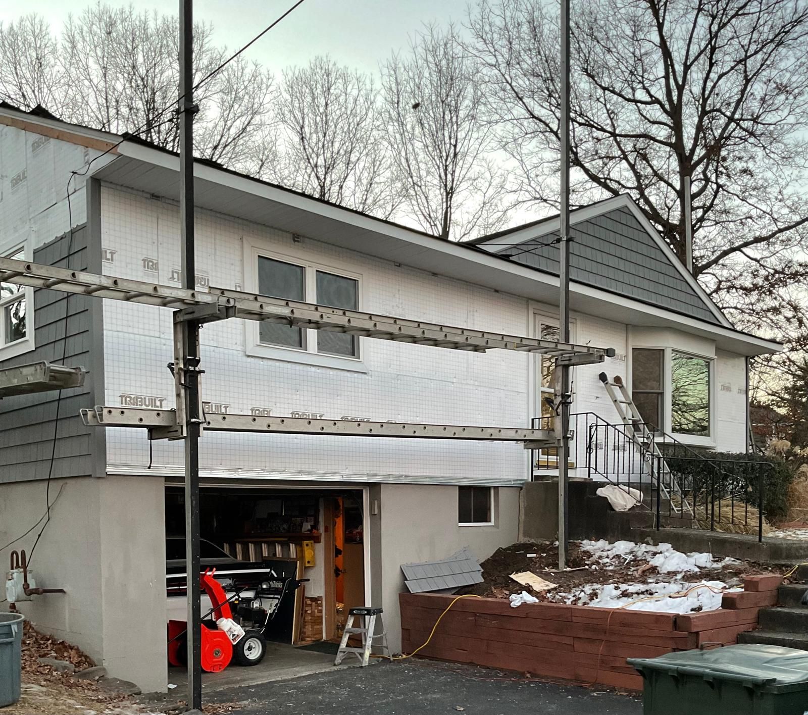 A house under renovation with scaffolding in front, showing exposed white house wrap and partial gray siding.