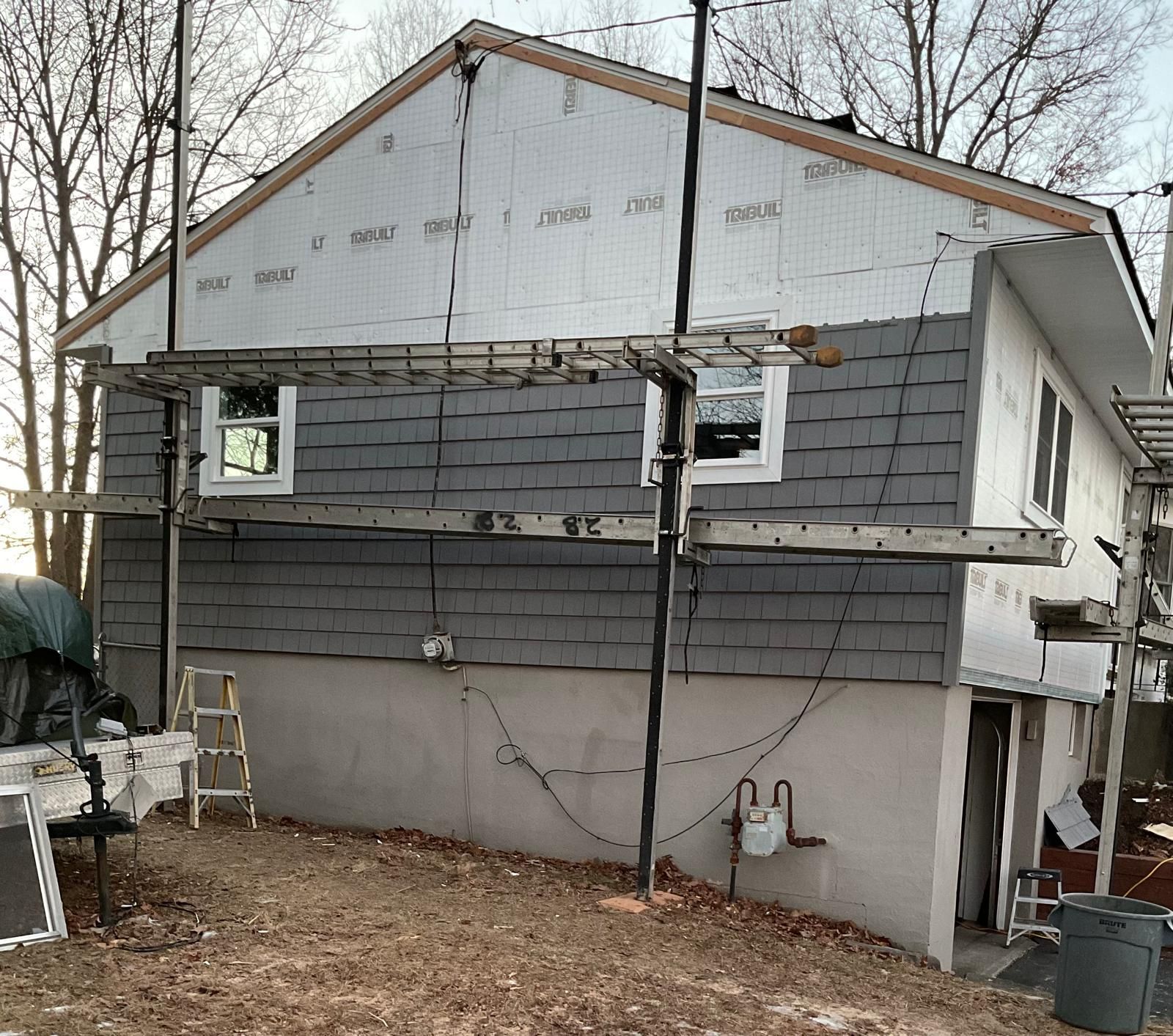 House exterior under renovation with grey siding, white insulation board, and metal scaffolding set up in the yard.