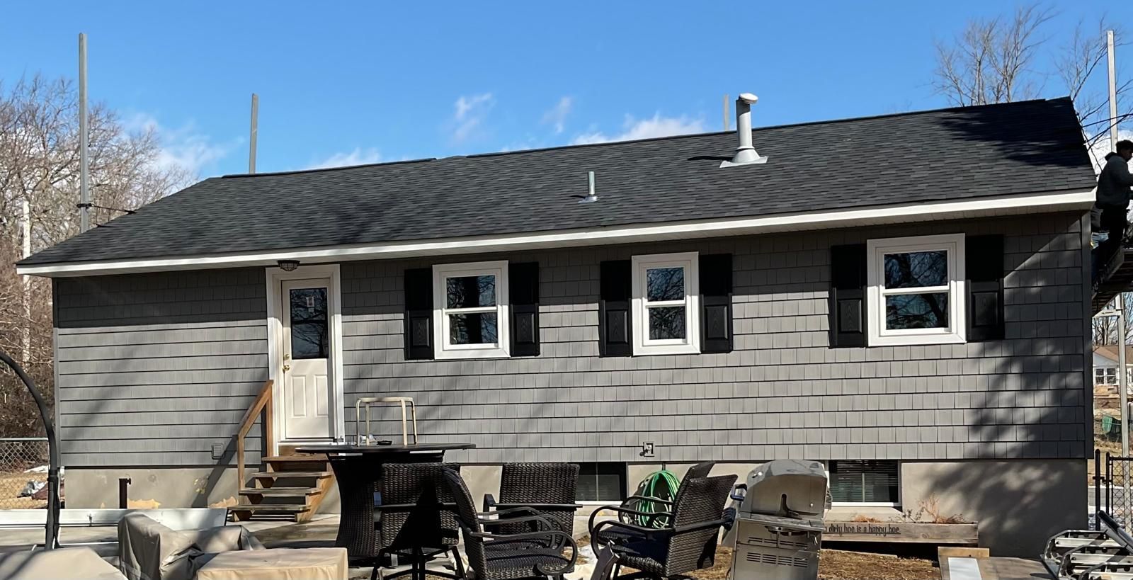A side view of a gray, single-story house with a black shingled roof, three windows with black shutters, and a back door.
