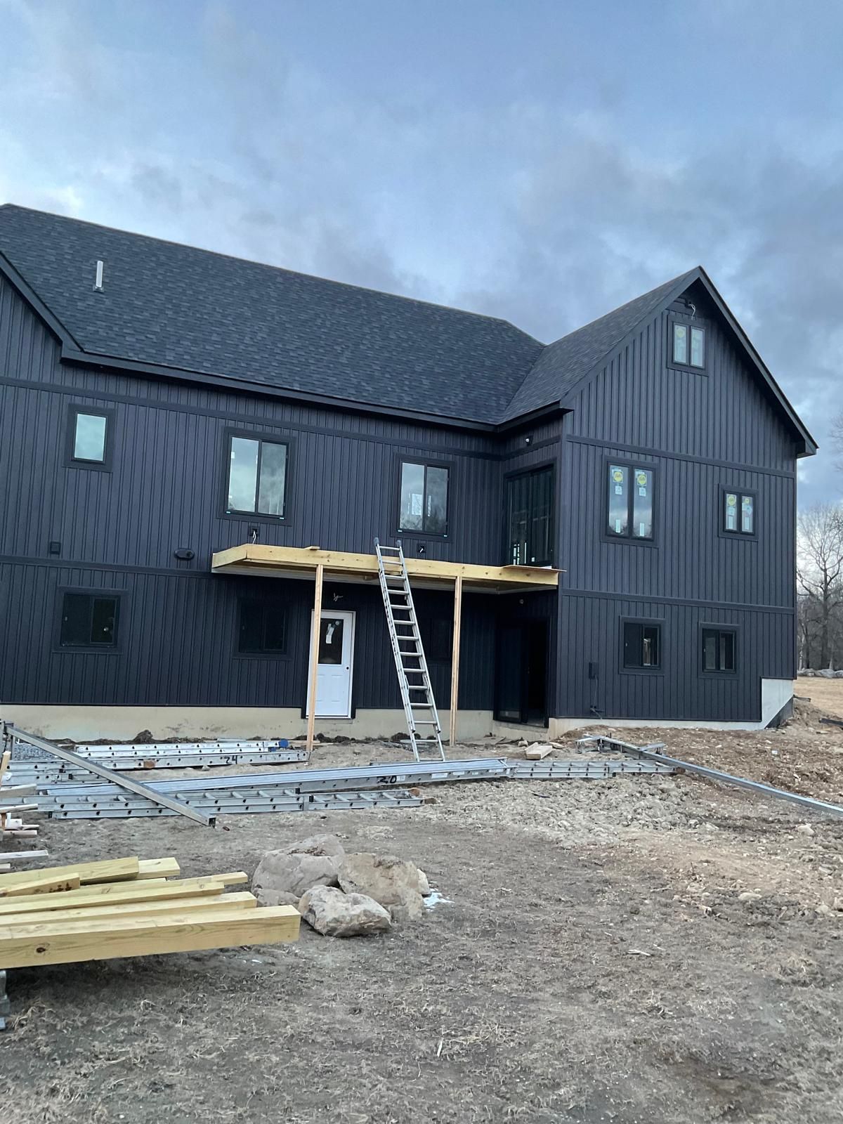 A modern black two-story home with vertical siding under construction, featuring a partially built wooden deck and ladder.