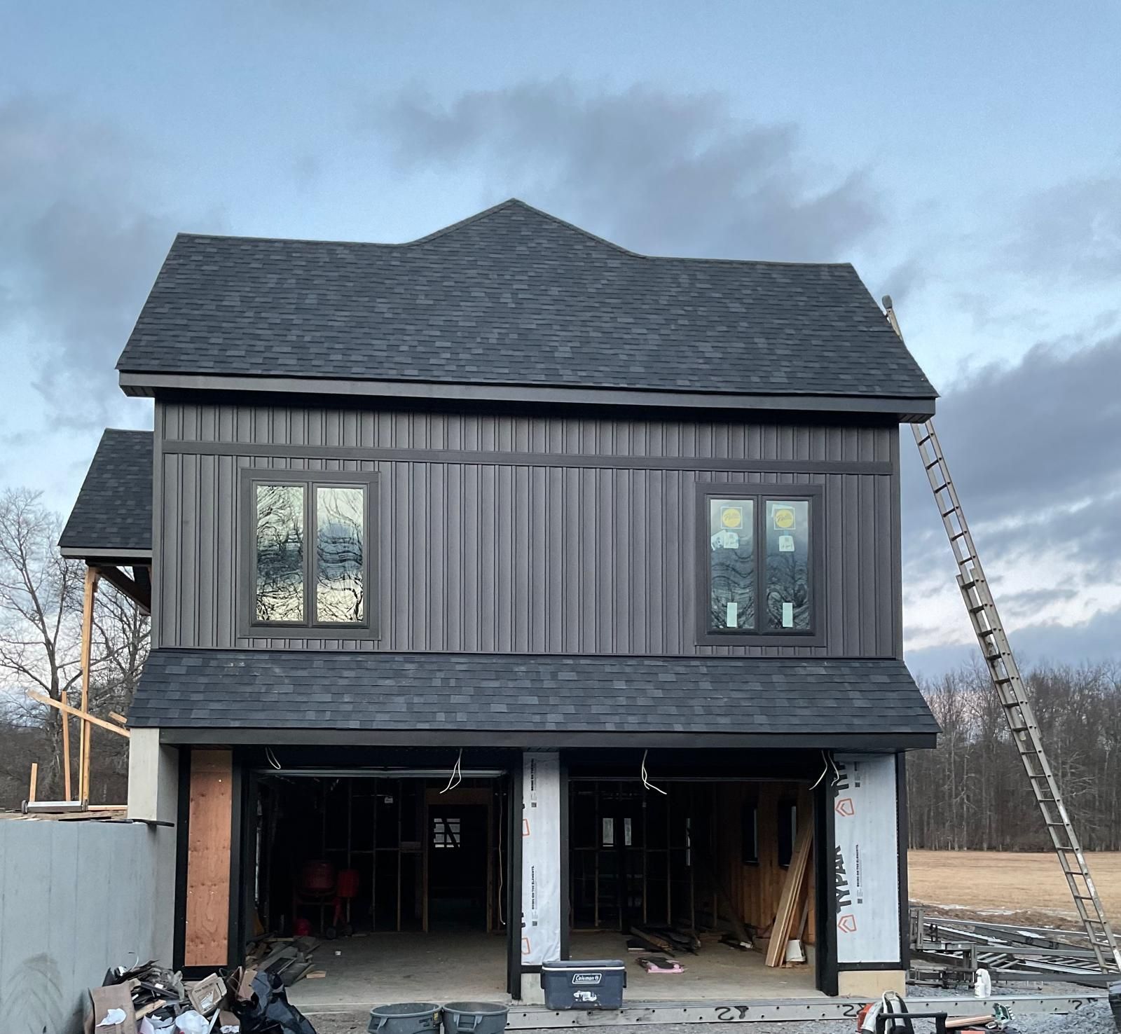 A two-story house under construction with dark vertical siding, a grey shingled roof, a double garage, and a tall ladder.