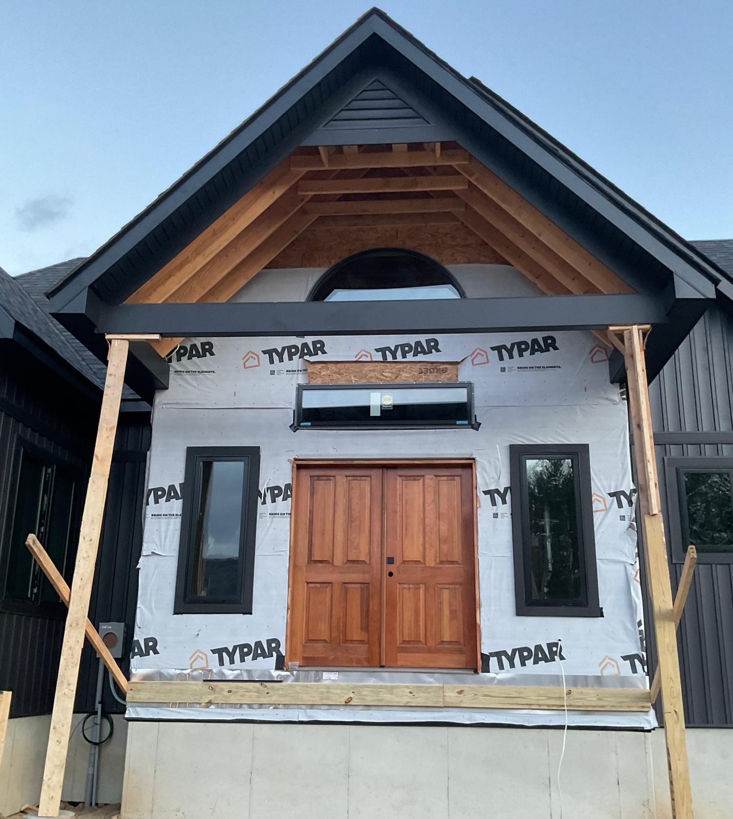 A modern house under construction featuring a wooden double door, arched window, and black trim under a gabled roof.