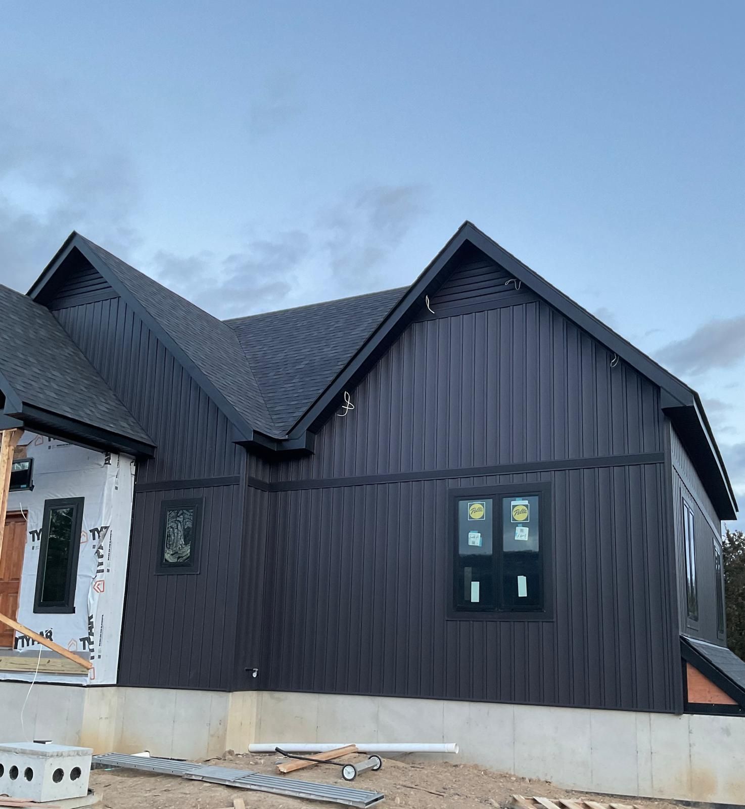 A newly constructed house with dark vertical siding, black-framed windows, and a shingled roof under a dusk sky.