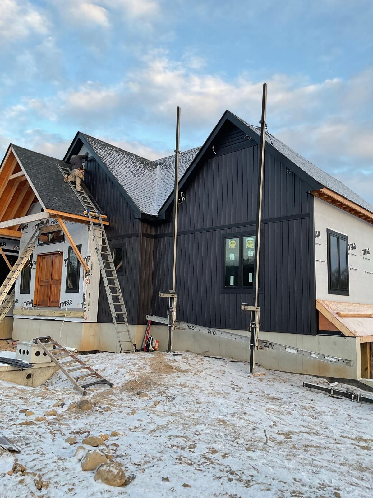 A house under construction with dark vertical siding, snow-covered ground, and wooden roof framing in a winter landscape.