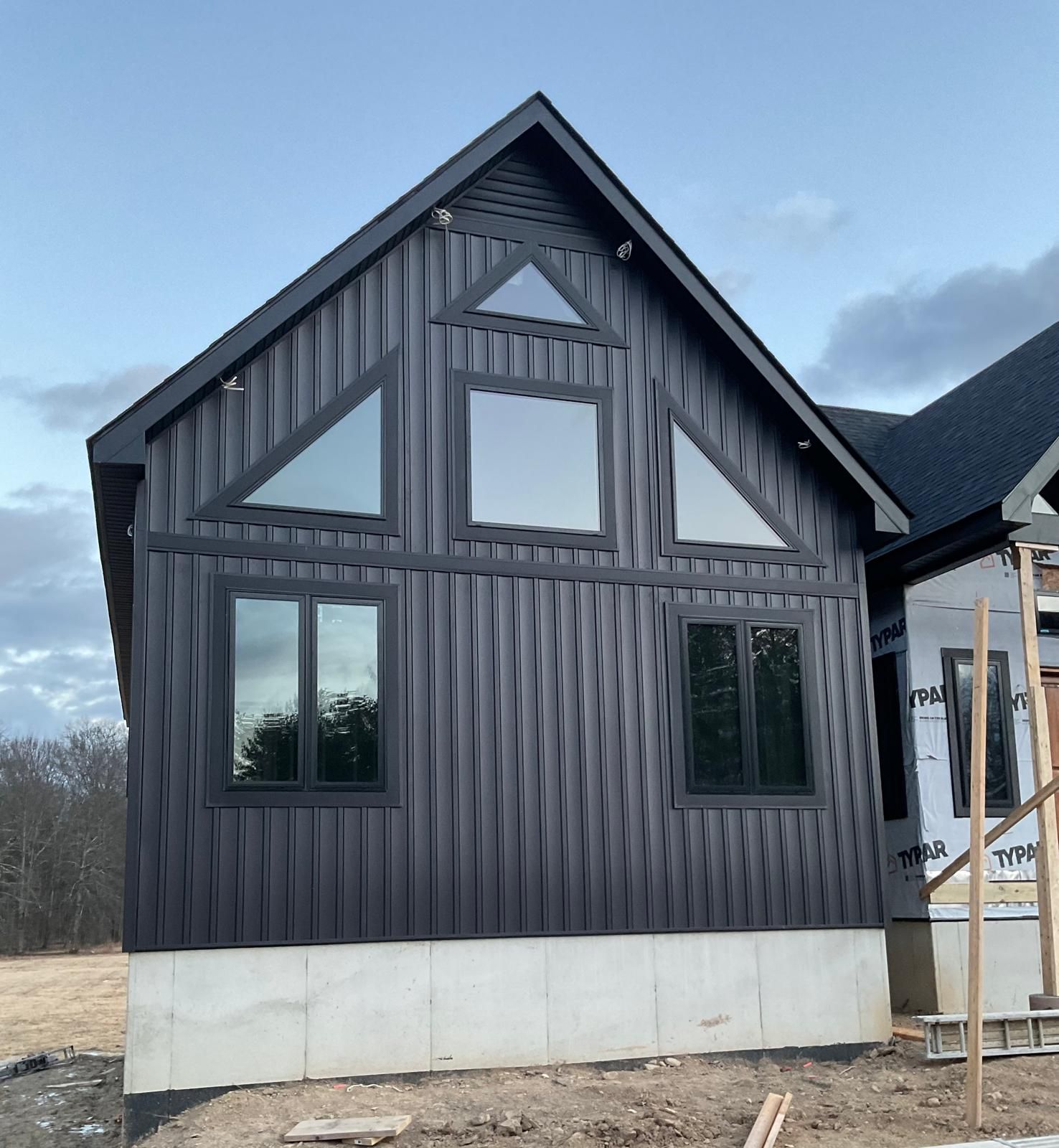 A modern house under construction featuring black vertical siding, large geometric windows, and a concrete foundation.