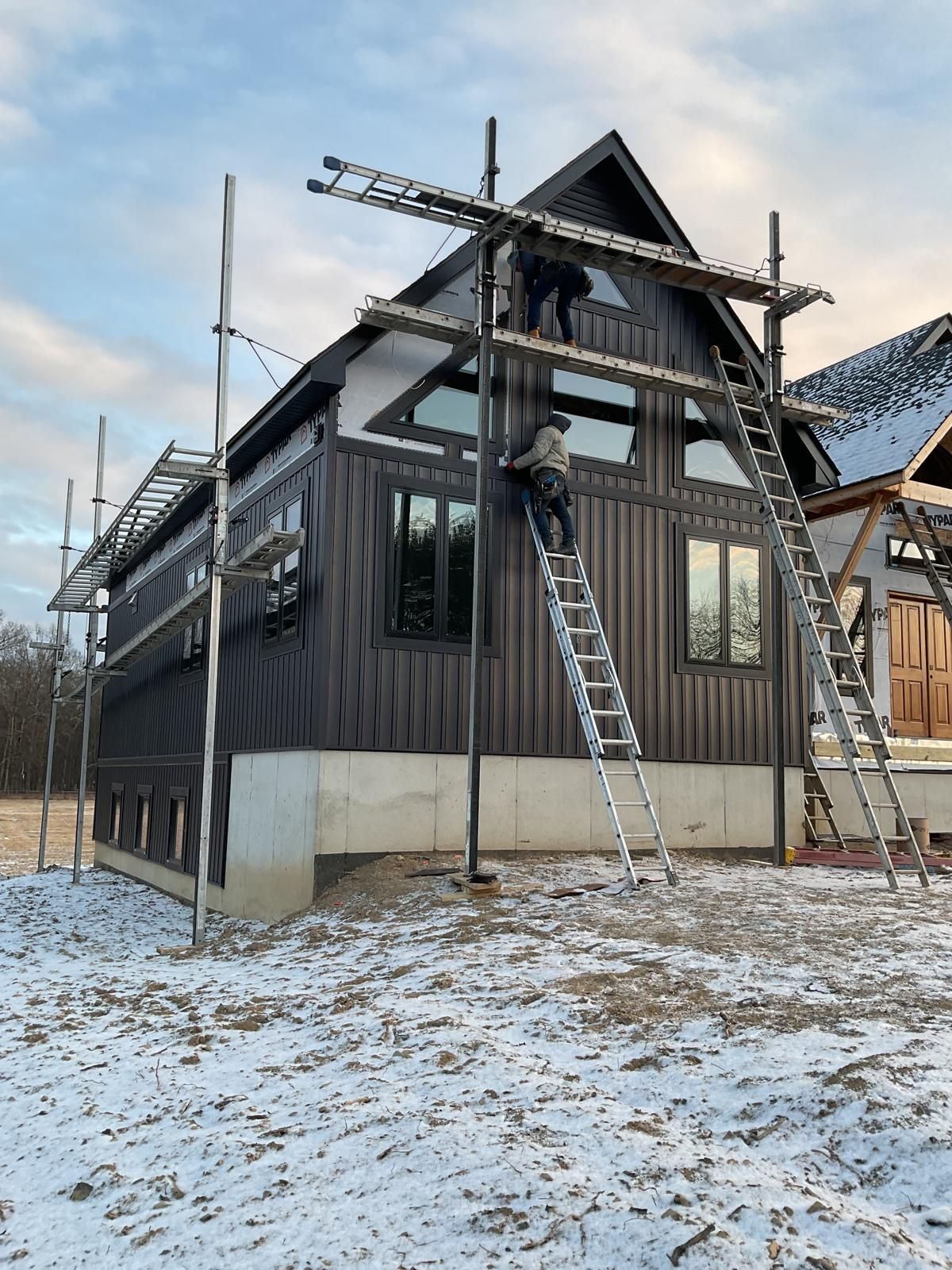Two construction workers on ladders install trim on a dark-sided modern house under construction in a snowy field.