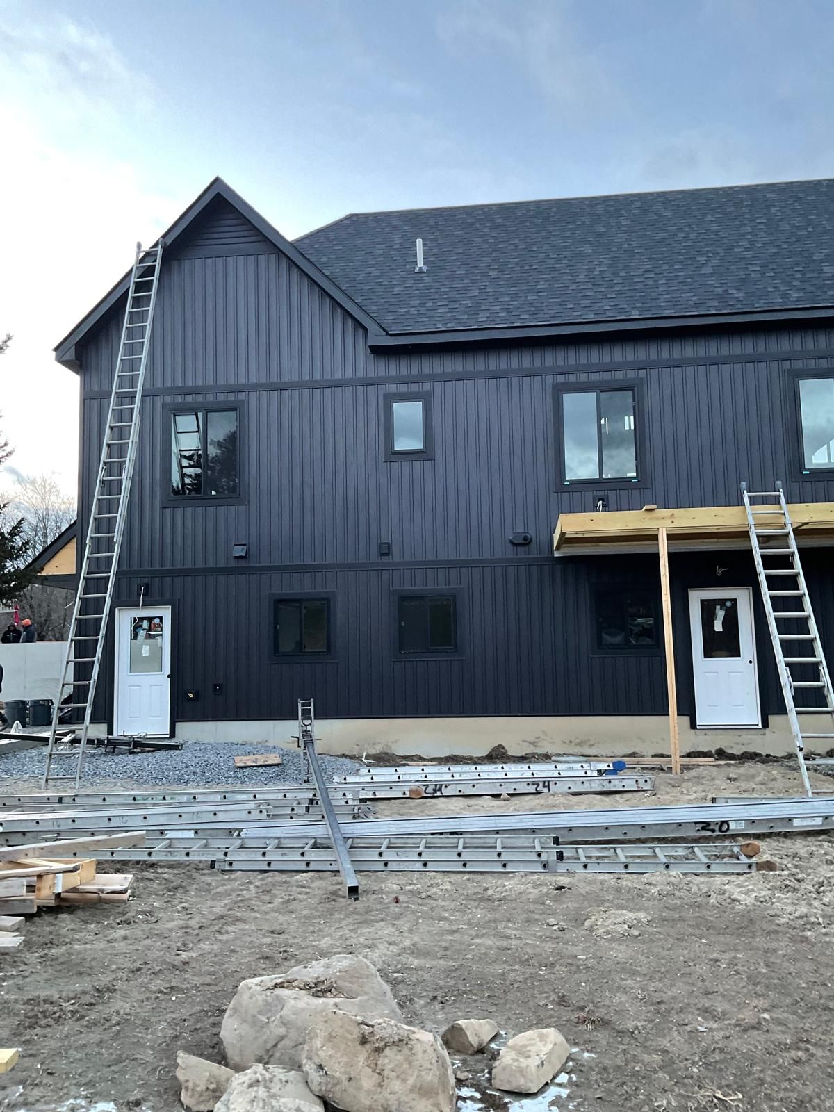 A two-story house under construction with dark vertical siding, a shingled roof, two white doors, and ladders.