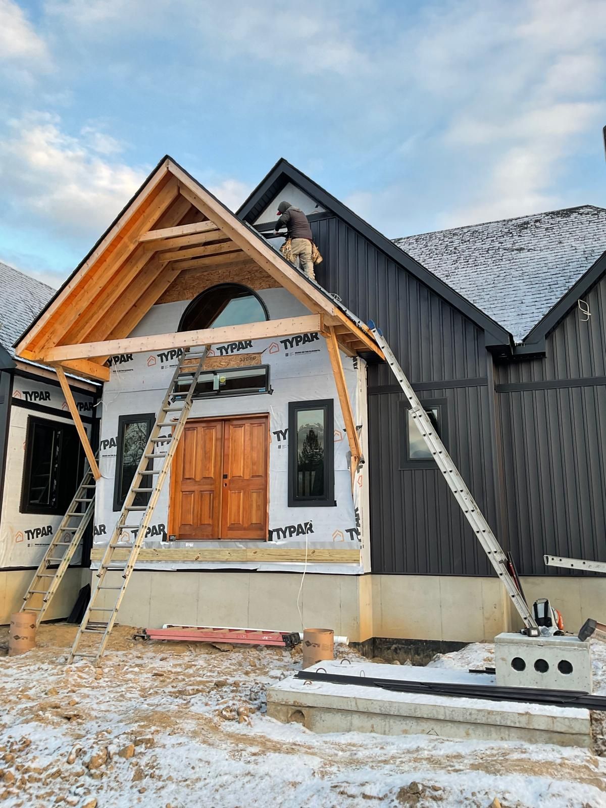A construction worker on a ladder installs wood trim on the peak of a modern home entryway under construction in snow.