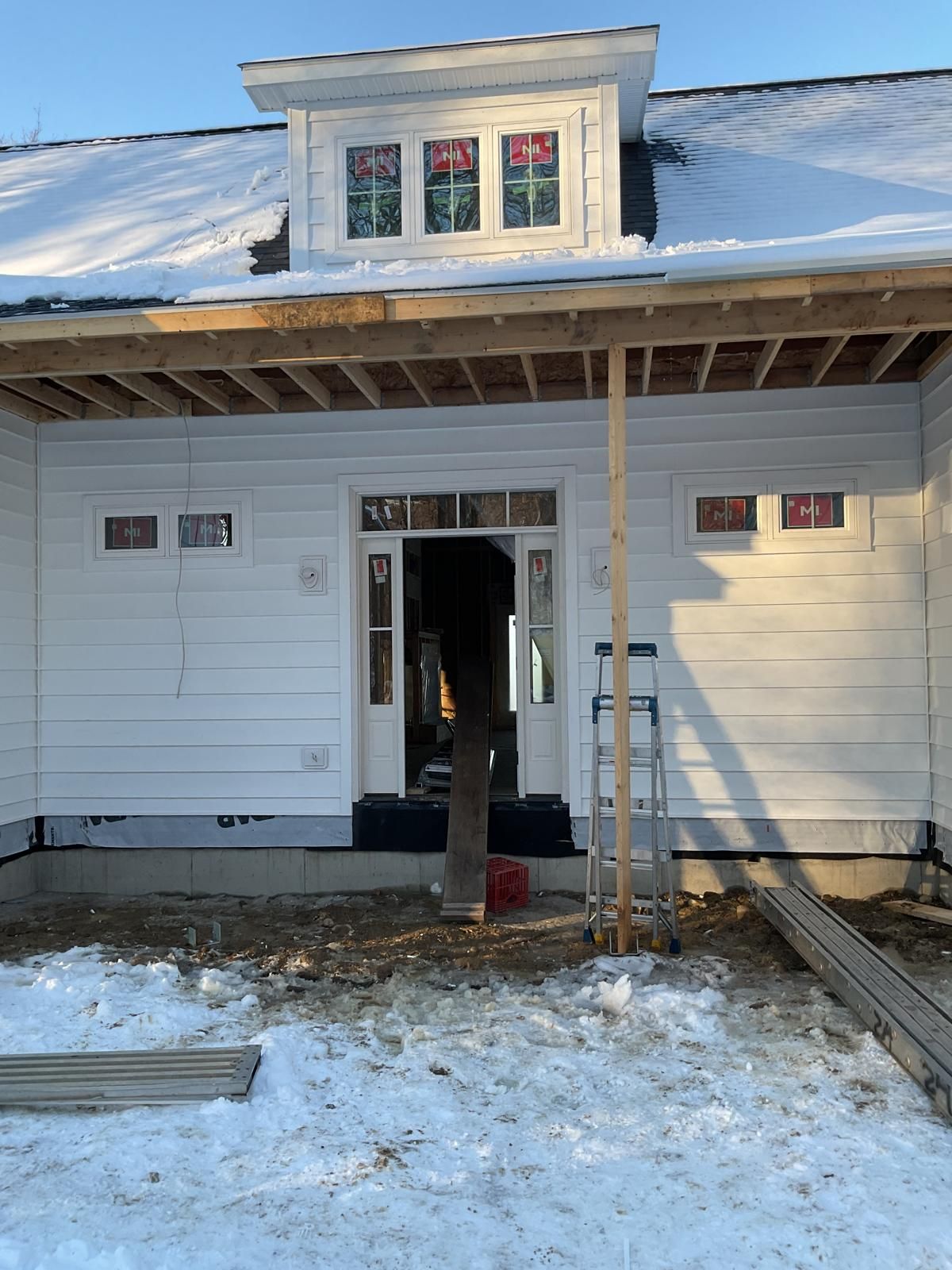 A partially constructed house exterior with white siding, a front entryway, a dormer window, and snow on the ground.