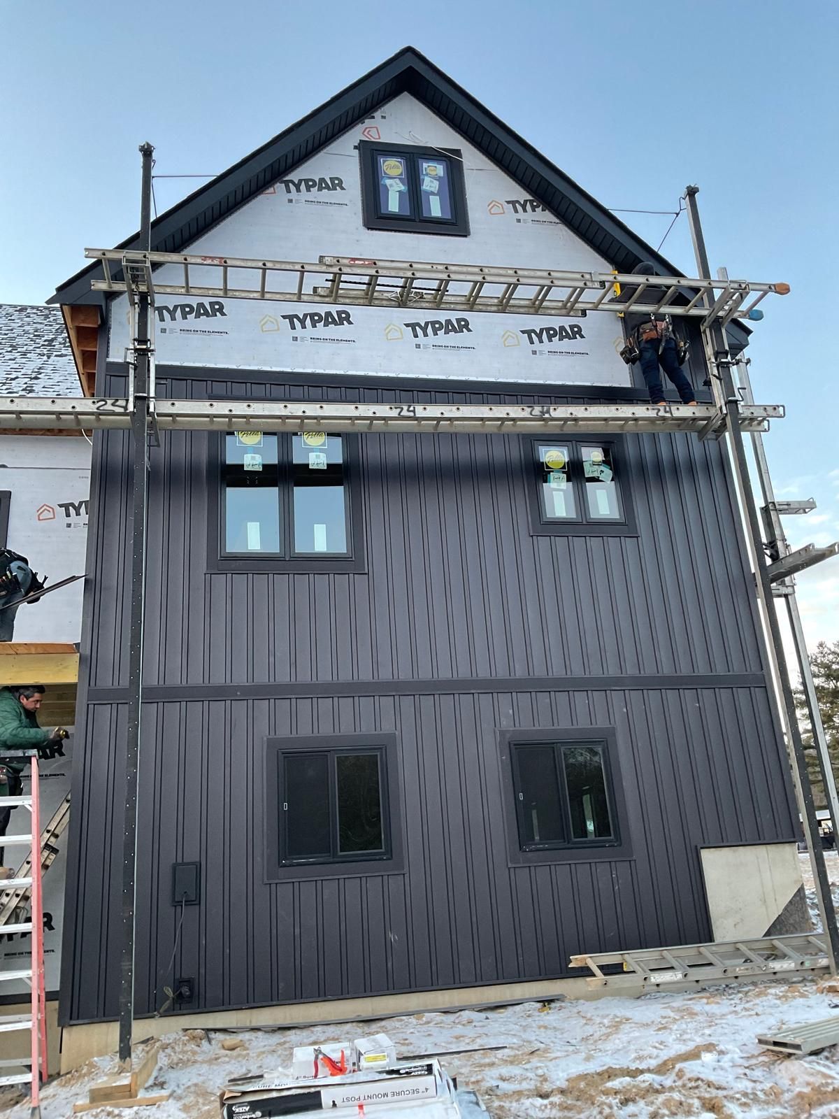 A two-story house under construction with dark vertical siding, scaffolding, and visible white weather-resistant wrap.