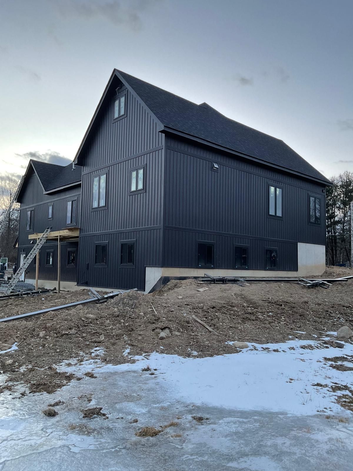 A two-story house with dark vertical siding under construction, surrounded by dirt and snow.