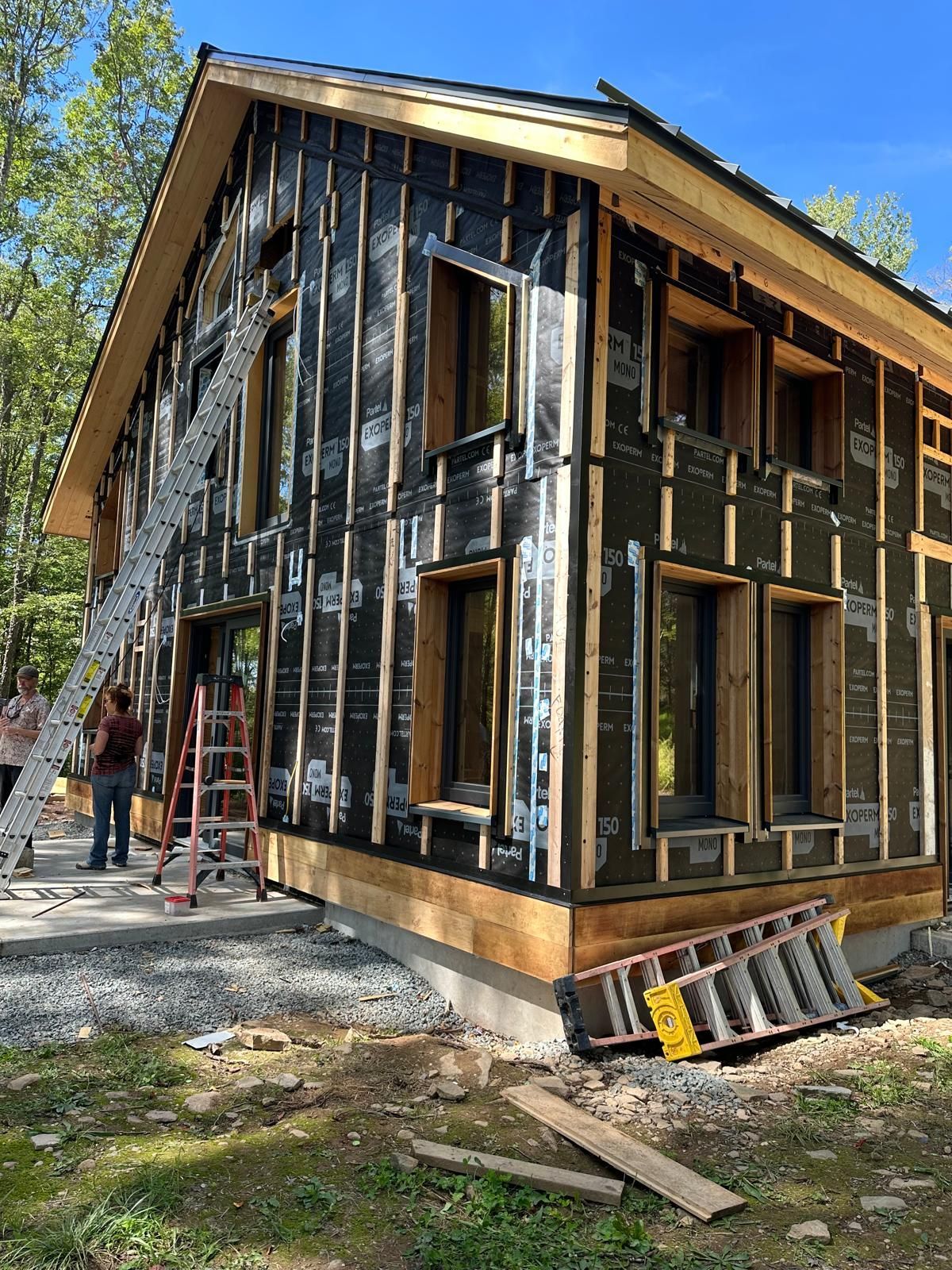 A partially constructed house under a blue sky, featuring black weatherproofing, wooden framing, and two ladders.