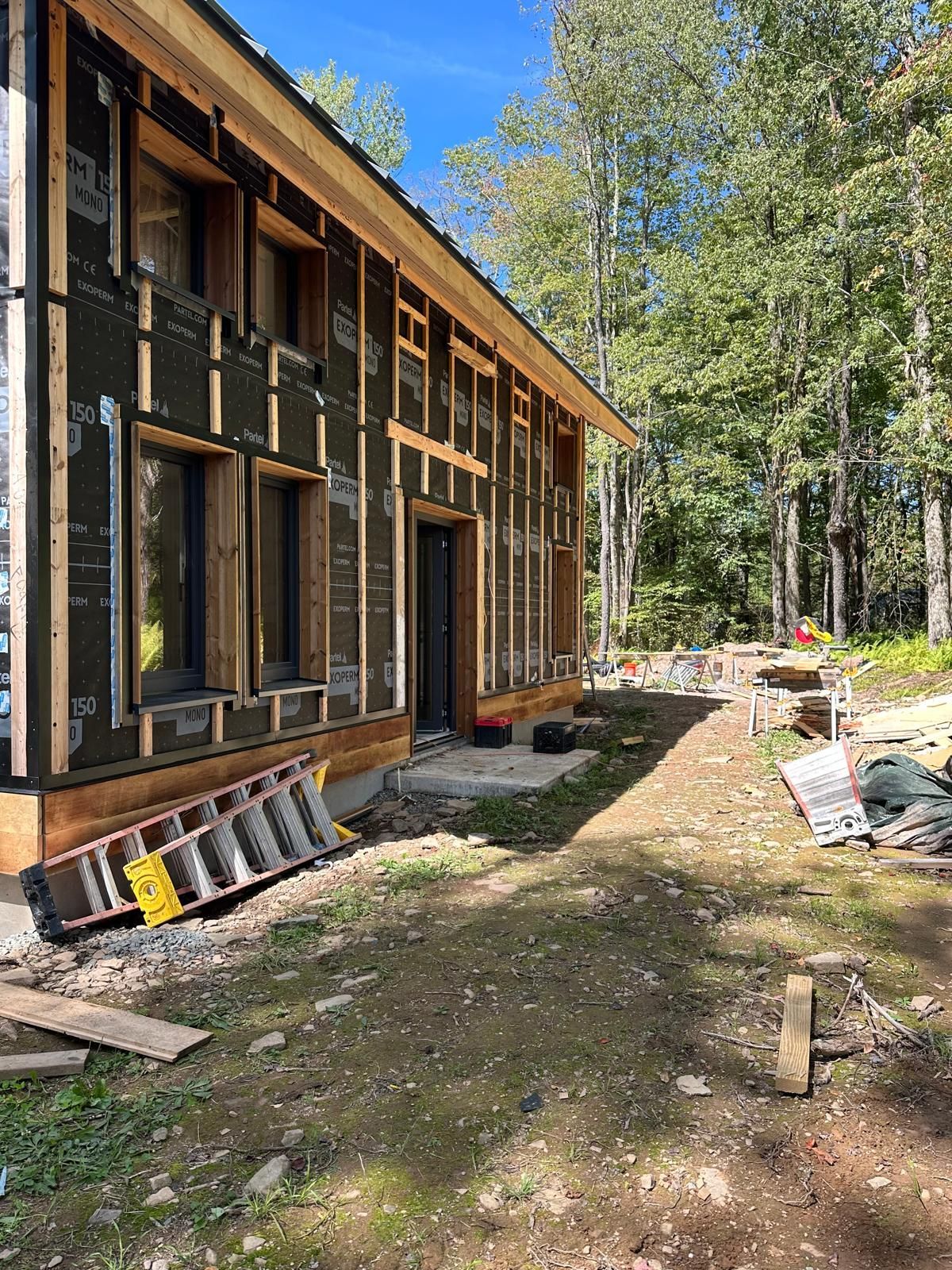 A wooden building under construction with exposed framing and black weather barrier, surrounded by a forest.