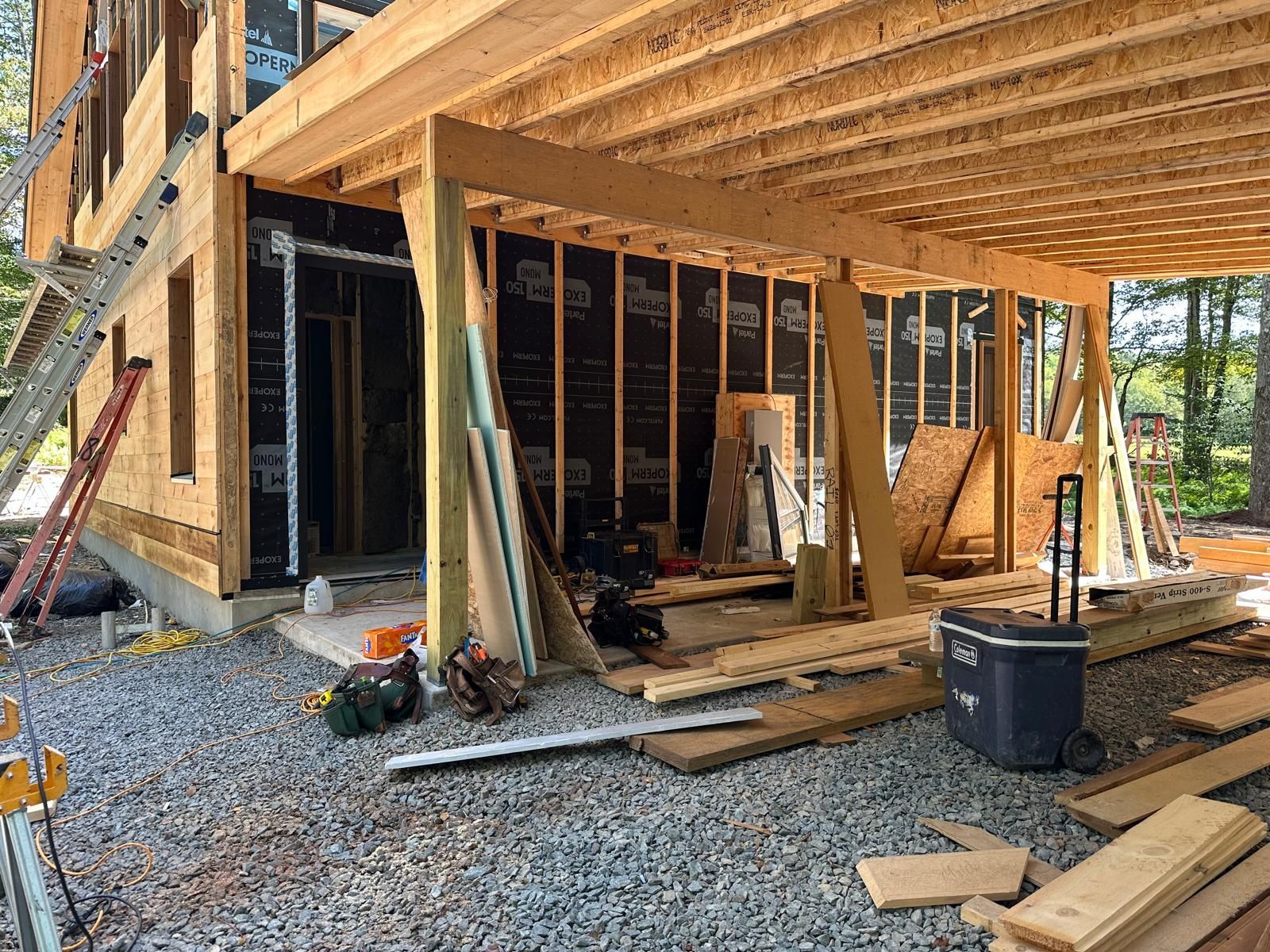 Construction site showing the timber frame and open floor joists of a new building, with a gravel ground and tools.