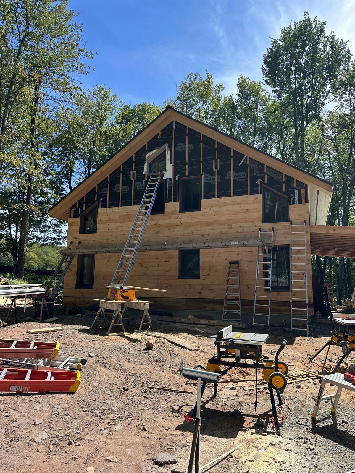A house under construction with exposed wooden siding and black sheathing, surrounded by scaffolding and power tools.
