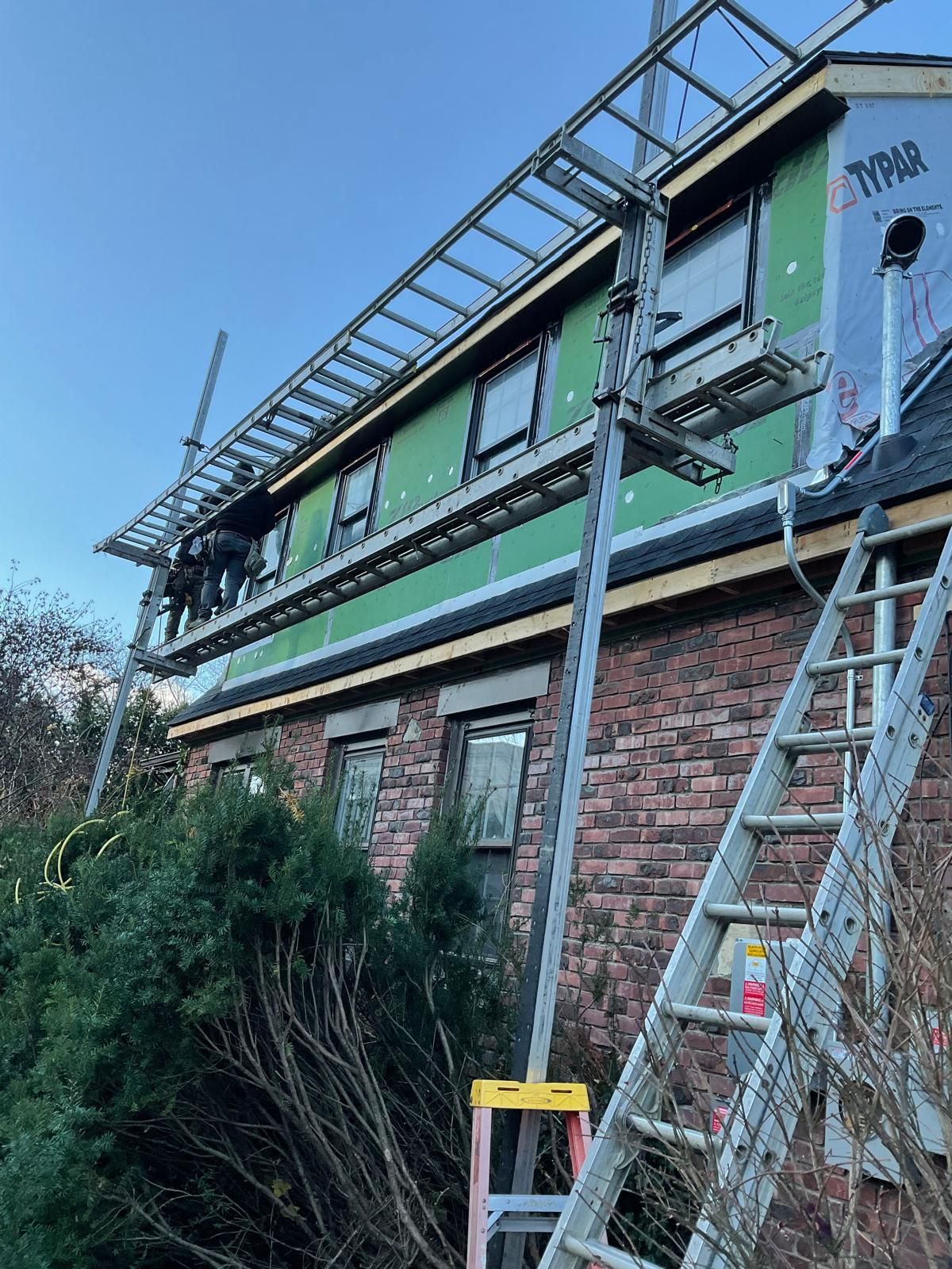 Workers on a metal pump jack scaffolding system installed against the side of a brick house with green exterior sheathing.