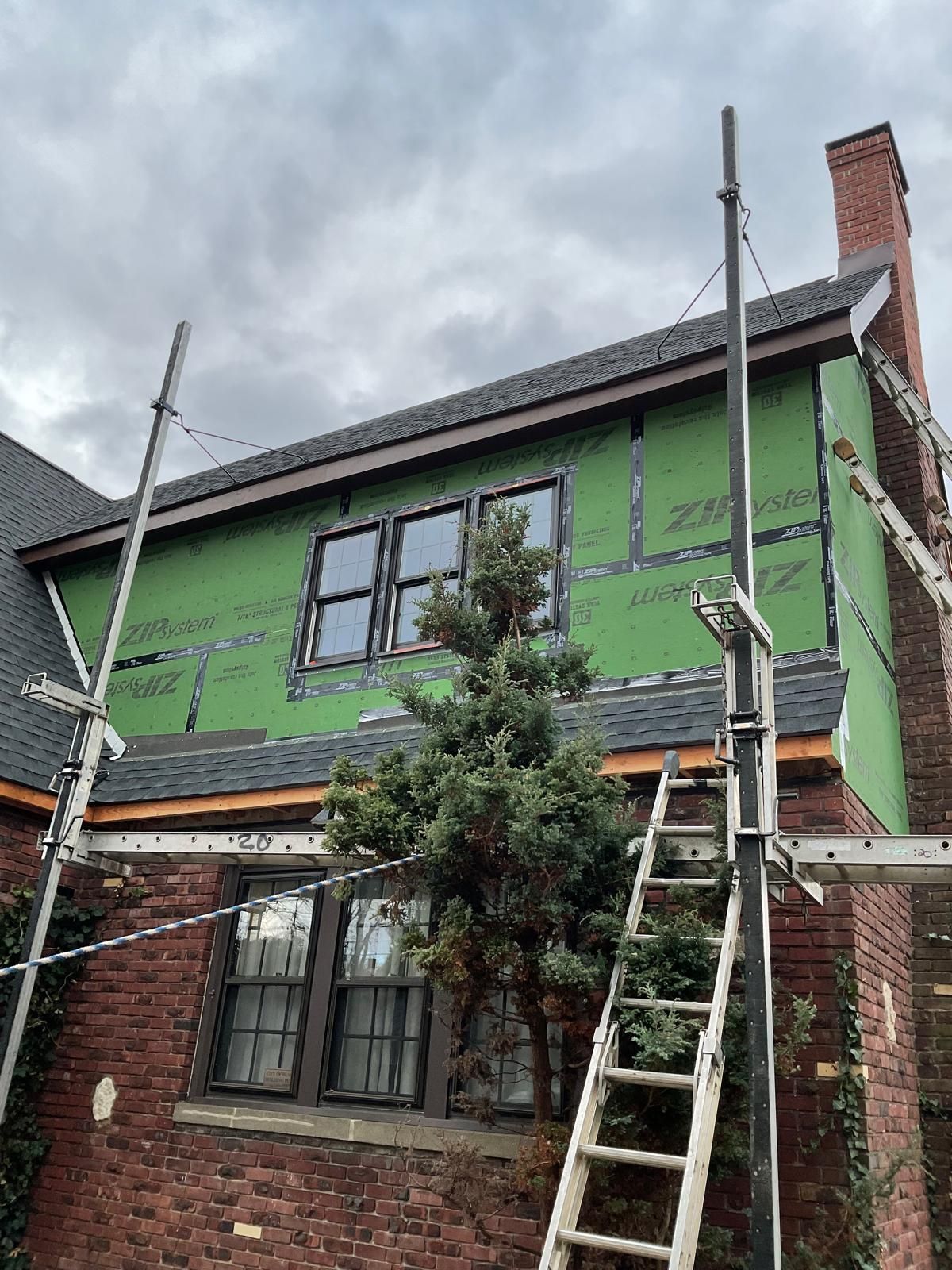 A red-brick house with green construction sheathing on the upper level, surrounded by scaffolding and a ladder.