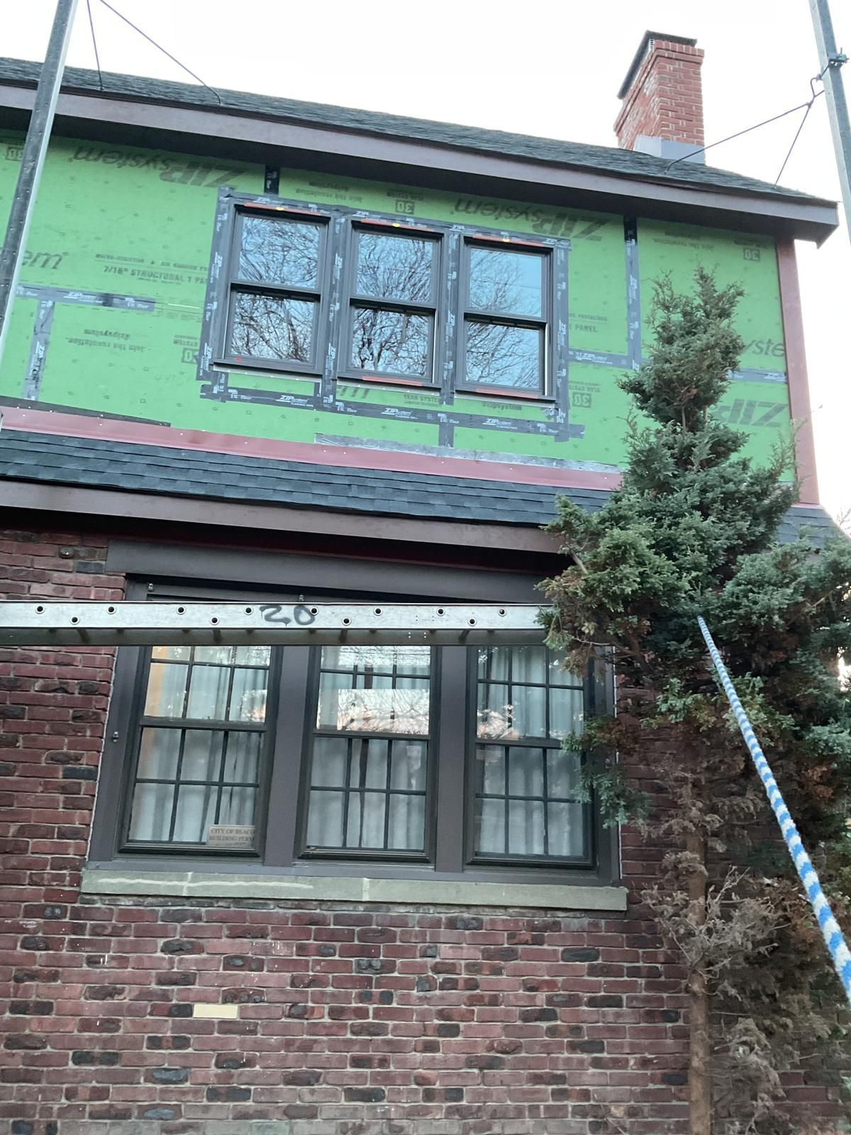 A two-story brick house under renovation with green structural sheathing on the upper level and a pine tree to the right.
