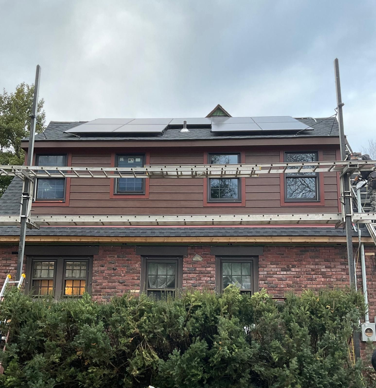 A two-story house with dark red siding, a brick lower level, and solar panels on the roof behind construction scaffolding.