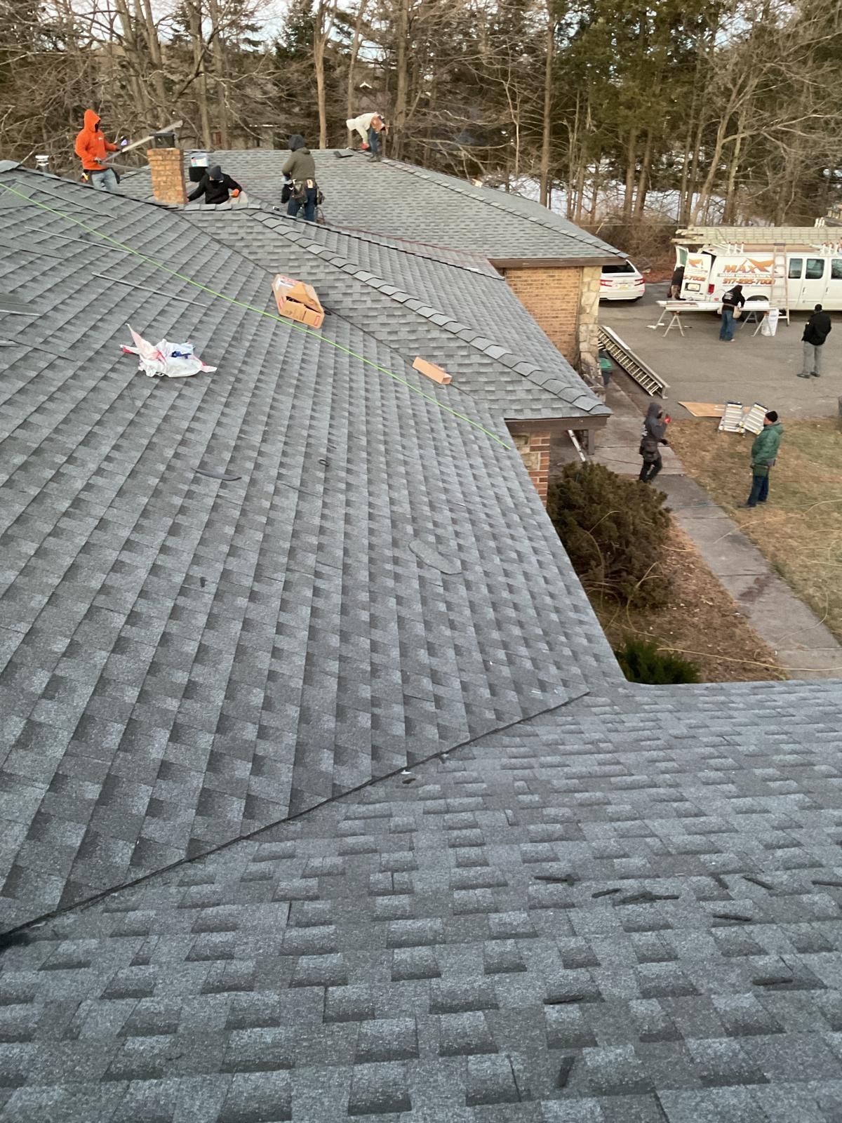 Workers in high-visibility gear install gray shingles on a residential roof, with others standing on the ground nearby.