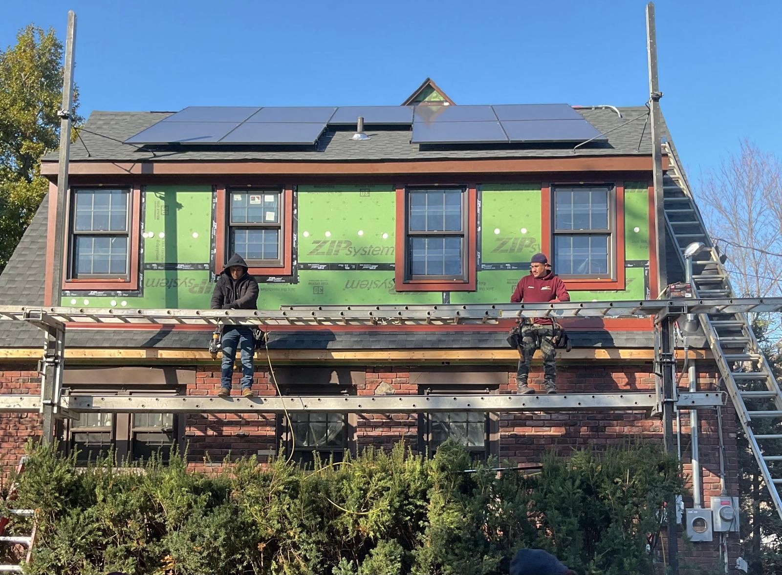 Two construction workers stand on scaffolding attached to a house with solar panels on the roof and green sheathing.
