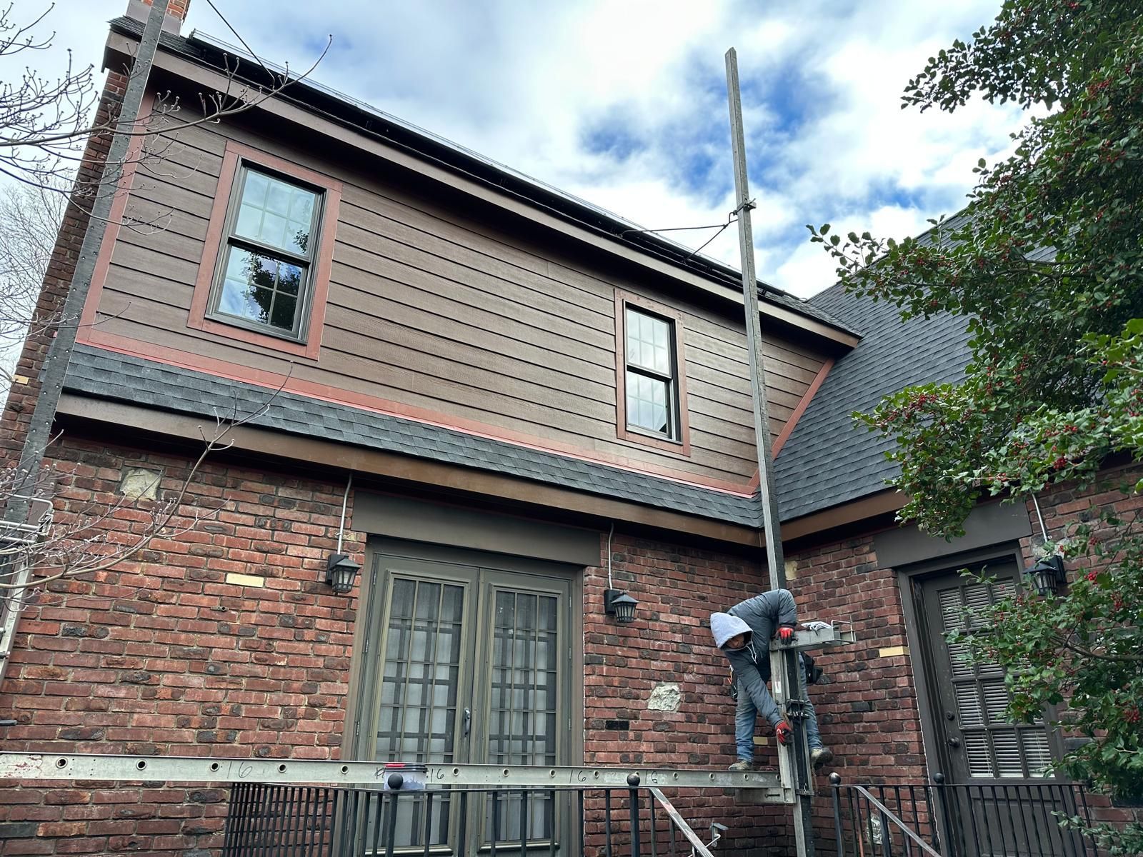 A person on a ladder repairs the exterior of a two-story brick house with a brown shingled upper level and glass doors.