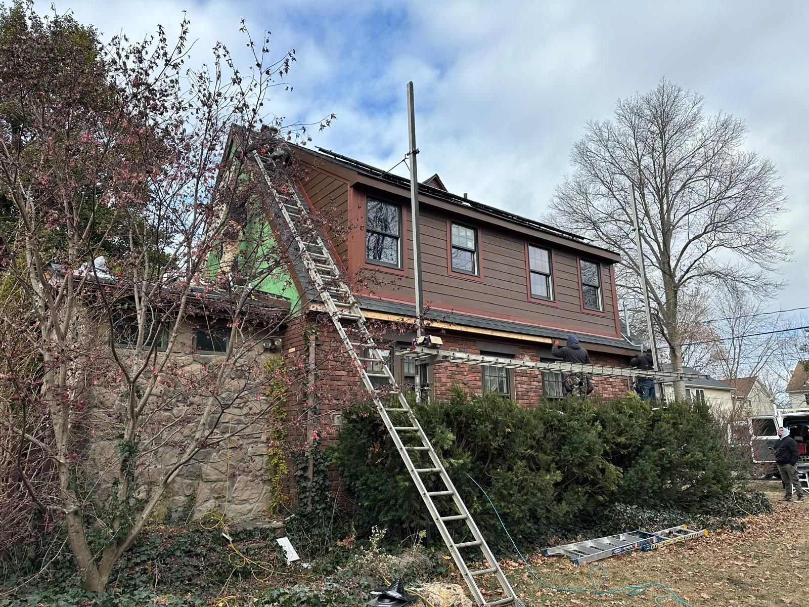 A ladder leans against a brick house undergoing exterior construction with visible insulation and brown roof shingles.