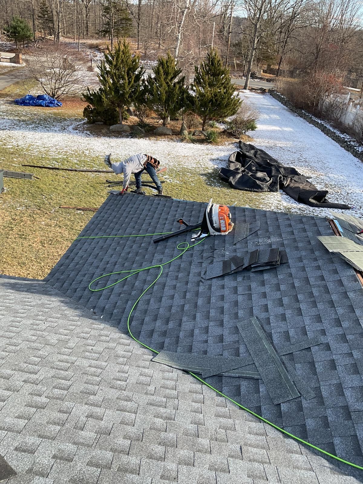A person works on a grey shingled roof while another person walks on the snowy ground below.