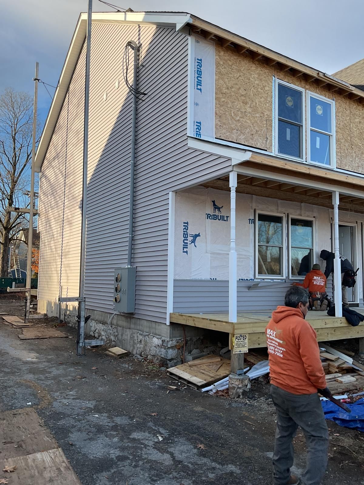 A person in an orange sweatshirt works on the porch of a house under construction with light gray siding and bare framing.