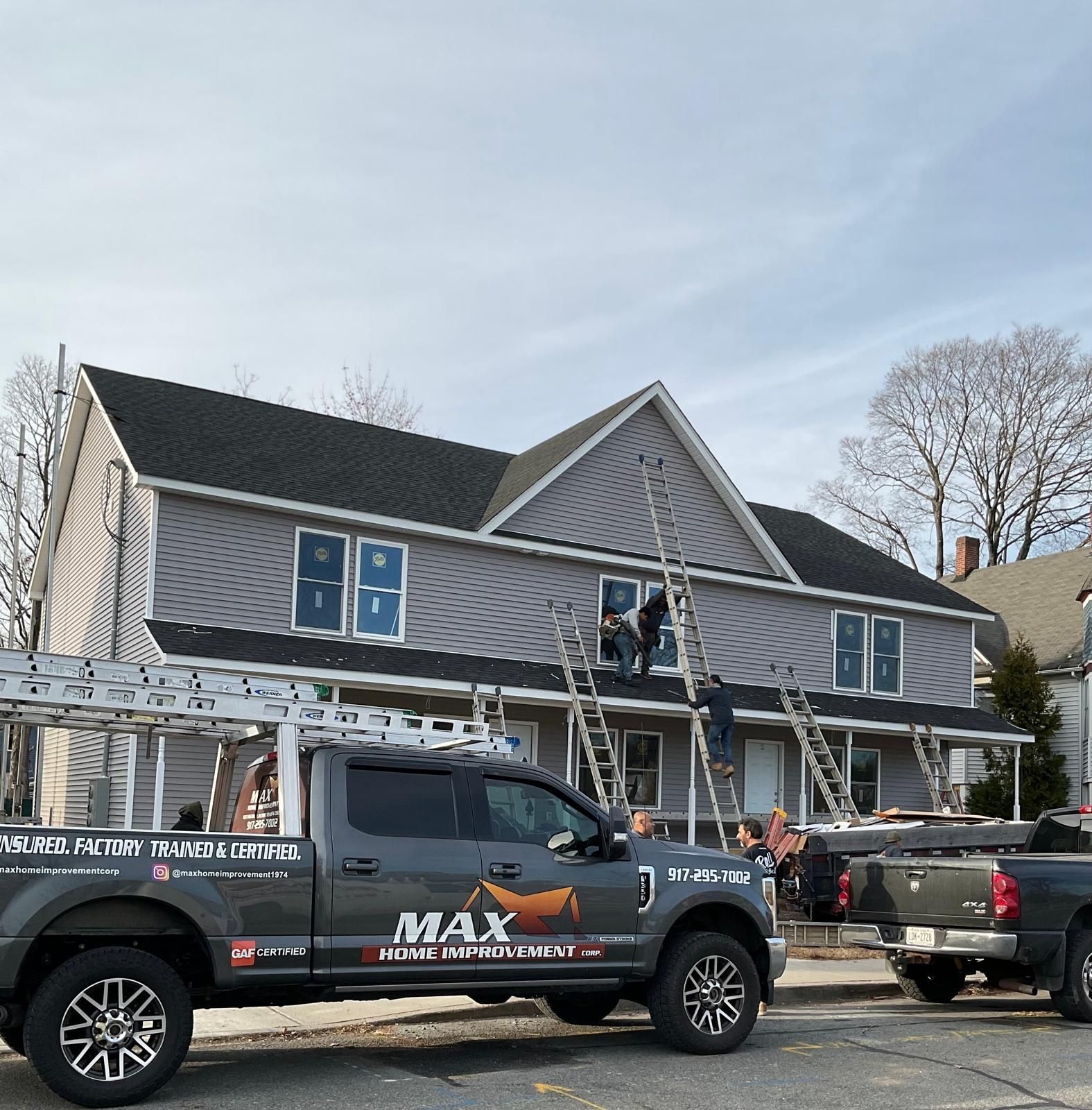 Workers use ladders to install siding on a gray, two-story house, with a Max Home Improvement truck parked in the foreground.