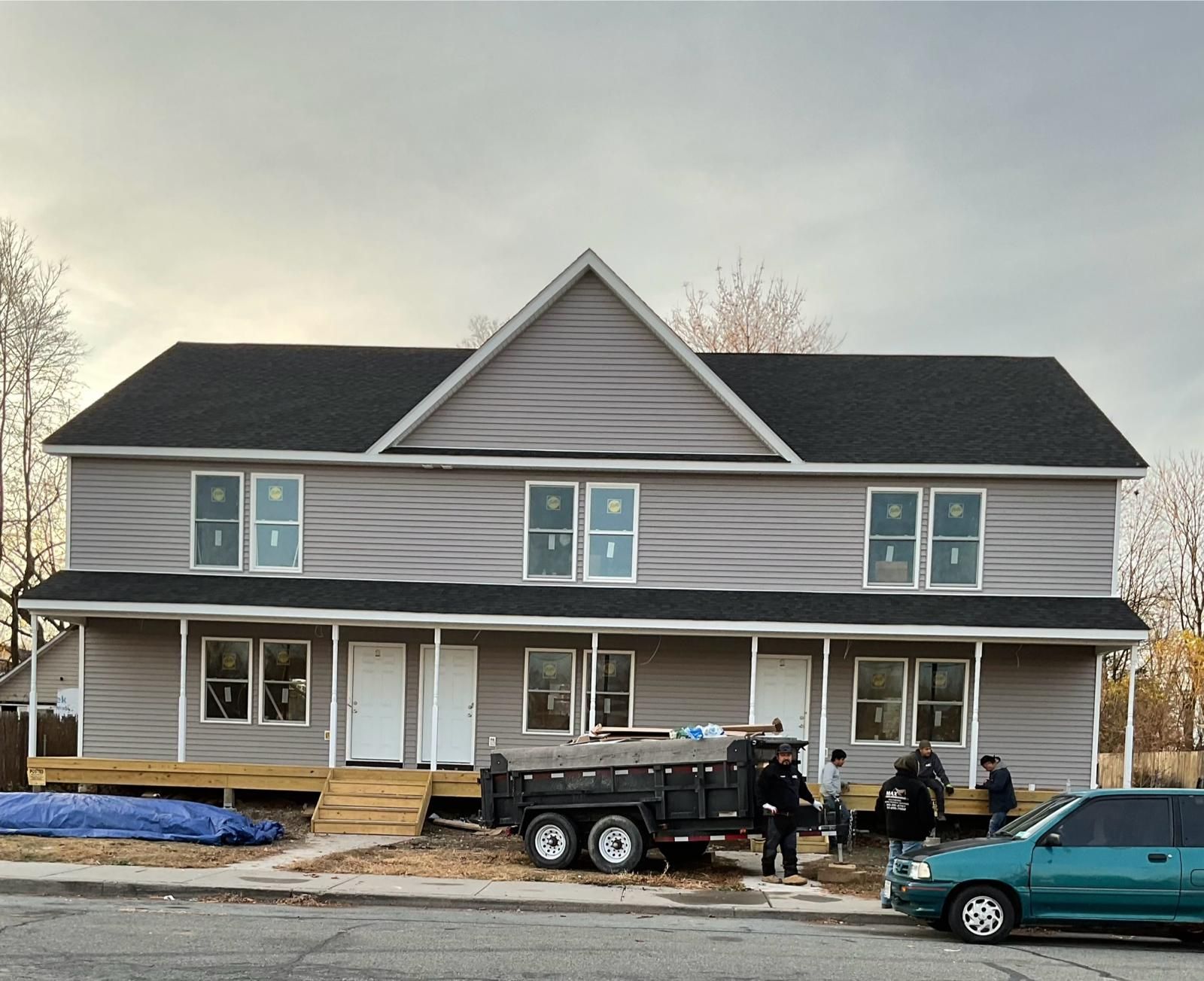 A two-story gray house under construction with a black roof, a wooden porch, and workers nearby.