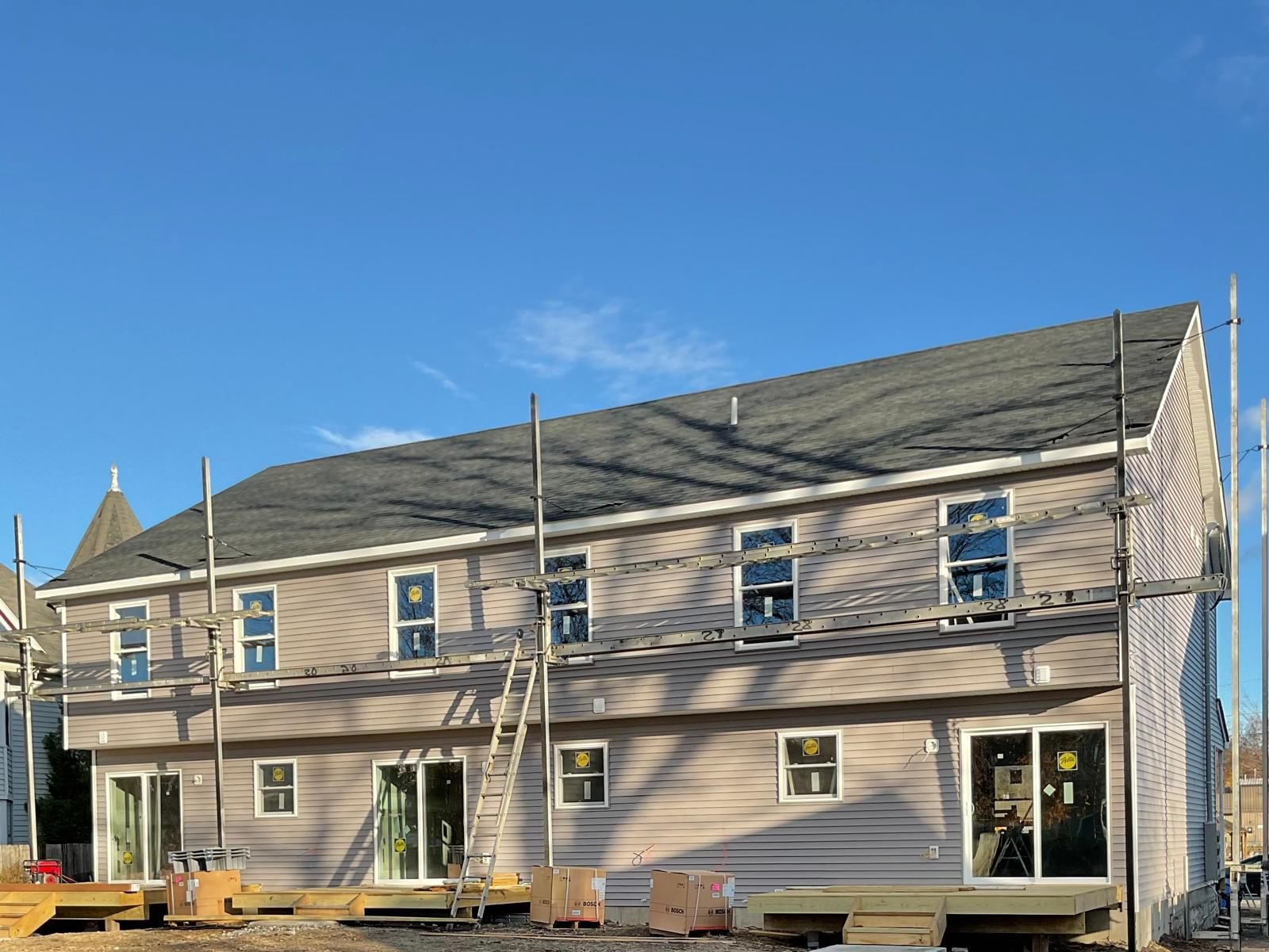 A two-story tan house under construction with scaffolding against its rear wall, visible sliding doors, and a dark roof.