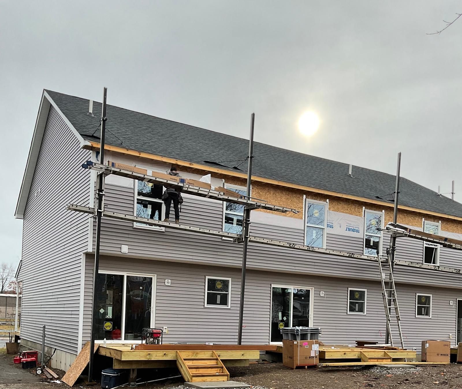 A two-story residential building under construction with light siding, a dark shingled roof, and unfinished wooden decks.
