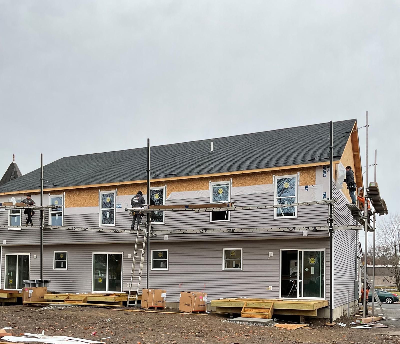 Workers on scaffolding install siding on a multi-story, dark-roofed residential building under construction.